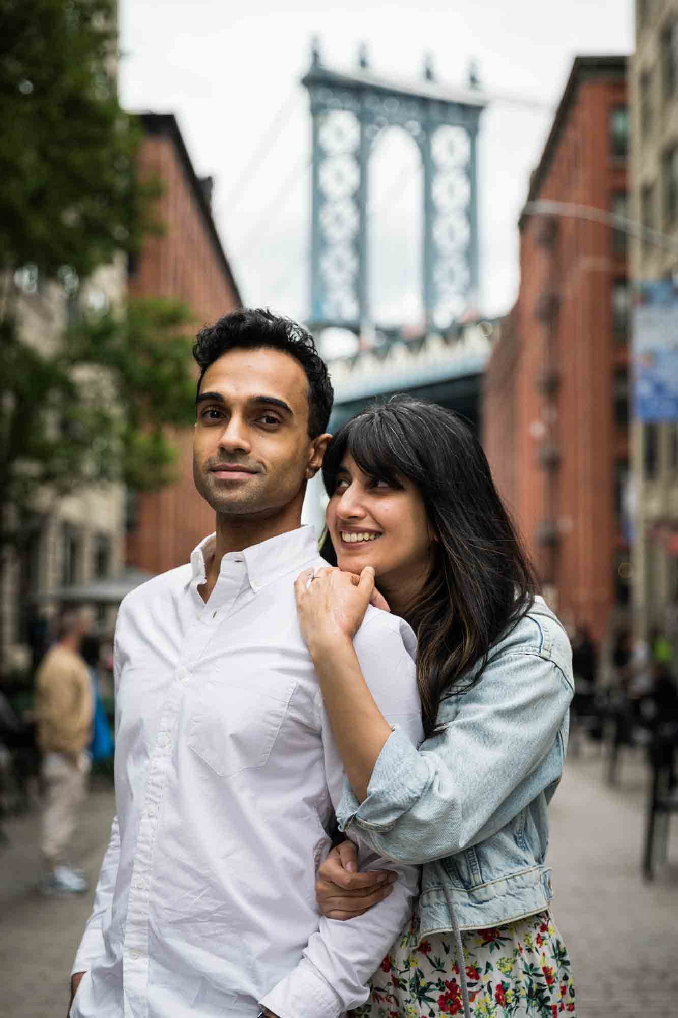 Woman standing behind man in front of Manhattan Bridge during a Brooklyn Bridge Park engagement photo shoot