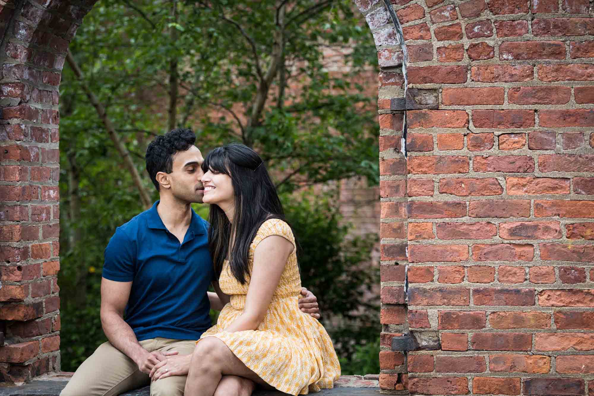 Brooklyn Bridge Park engagement photo of man kissing woman on cheek while sitting in brick window
