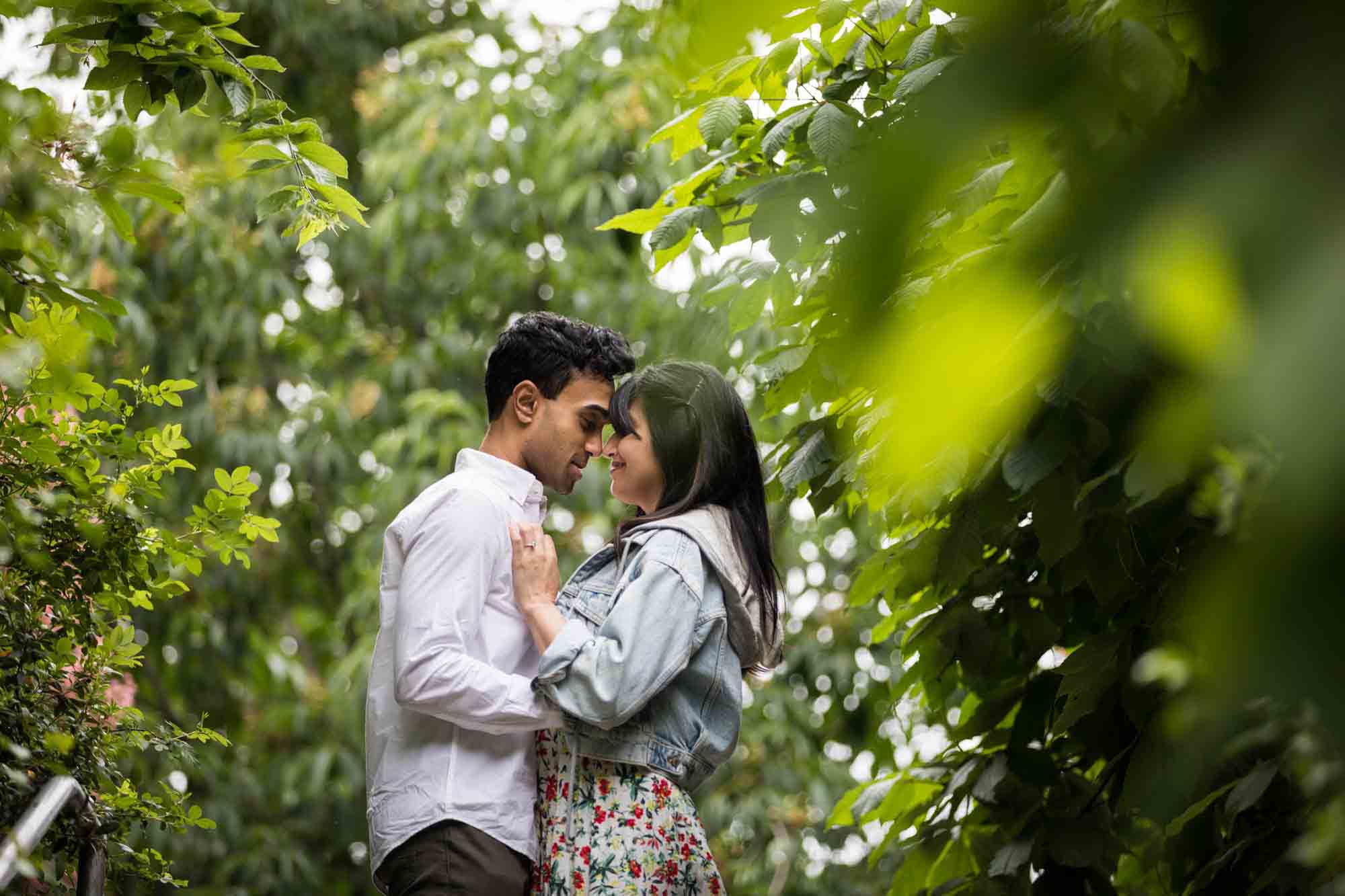 Couple cuddling in front of trees and bushes during a Brooklyn Bridge Park engagement photo shoot