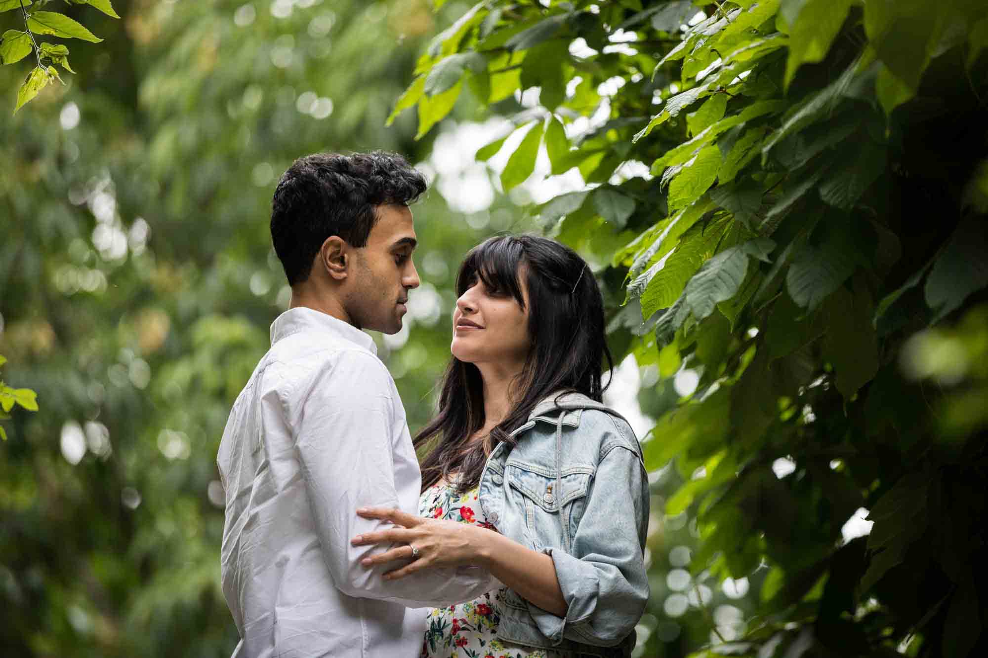 Couple cuddling in front of trees and plants during a Brooklyn Bridge Park engagement photo shoot