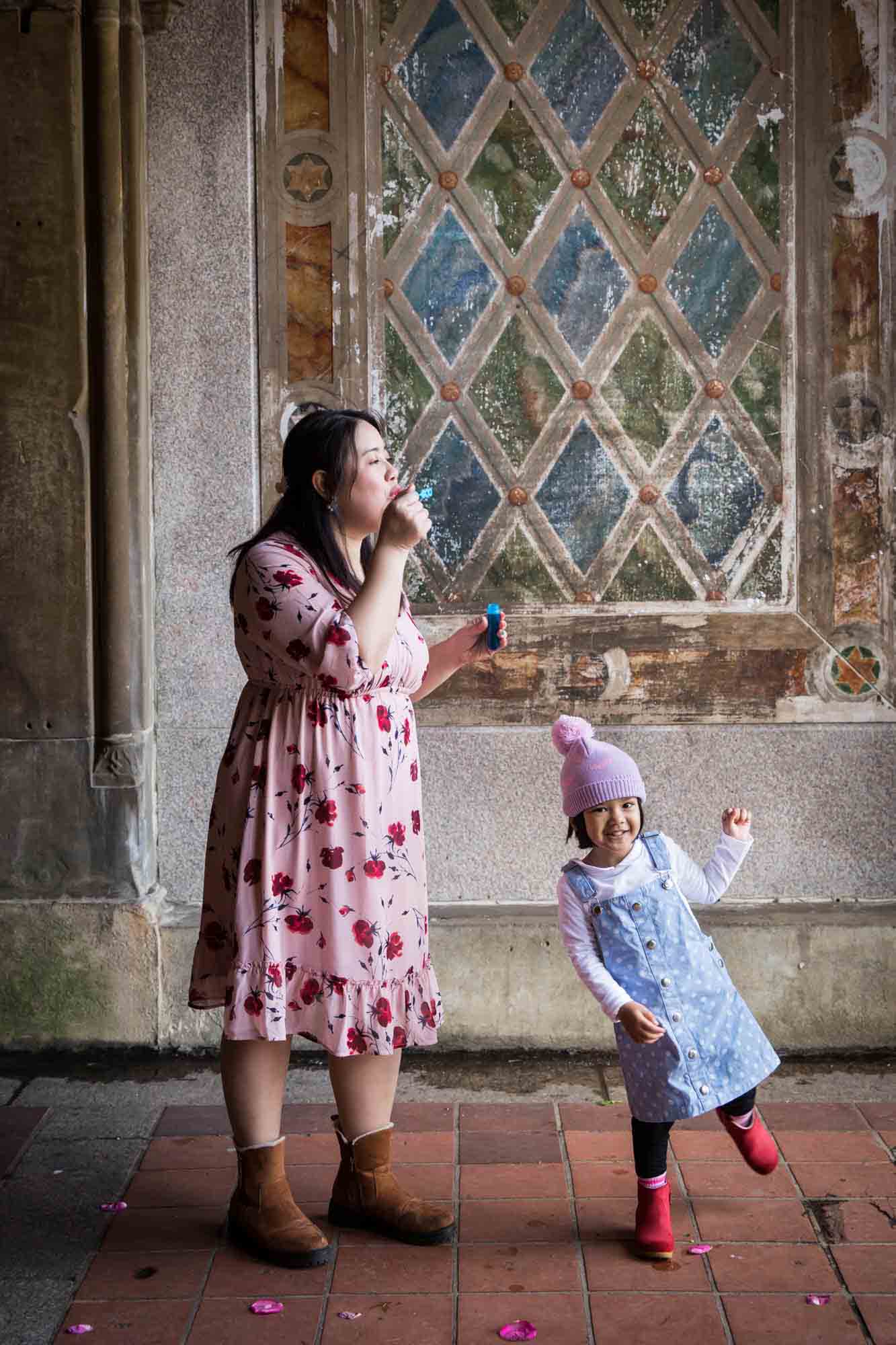 Mother blowing bubbles over little girl wearing knit cap during a Central Park family portrait