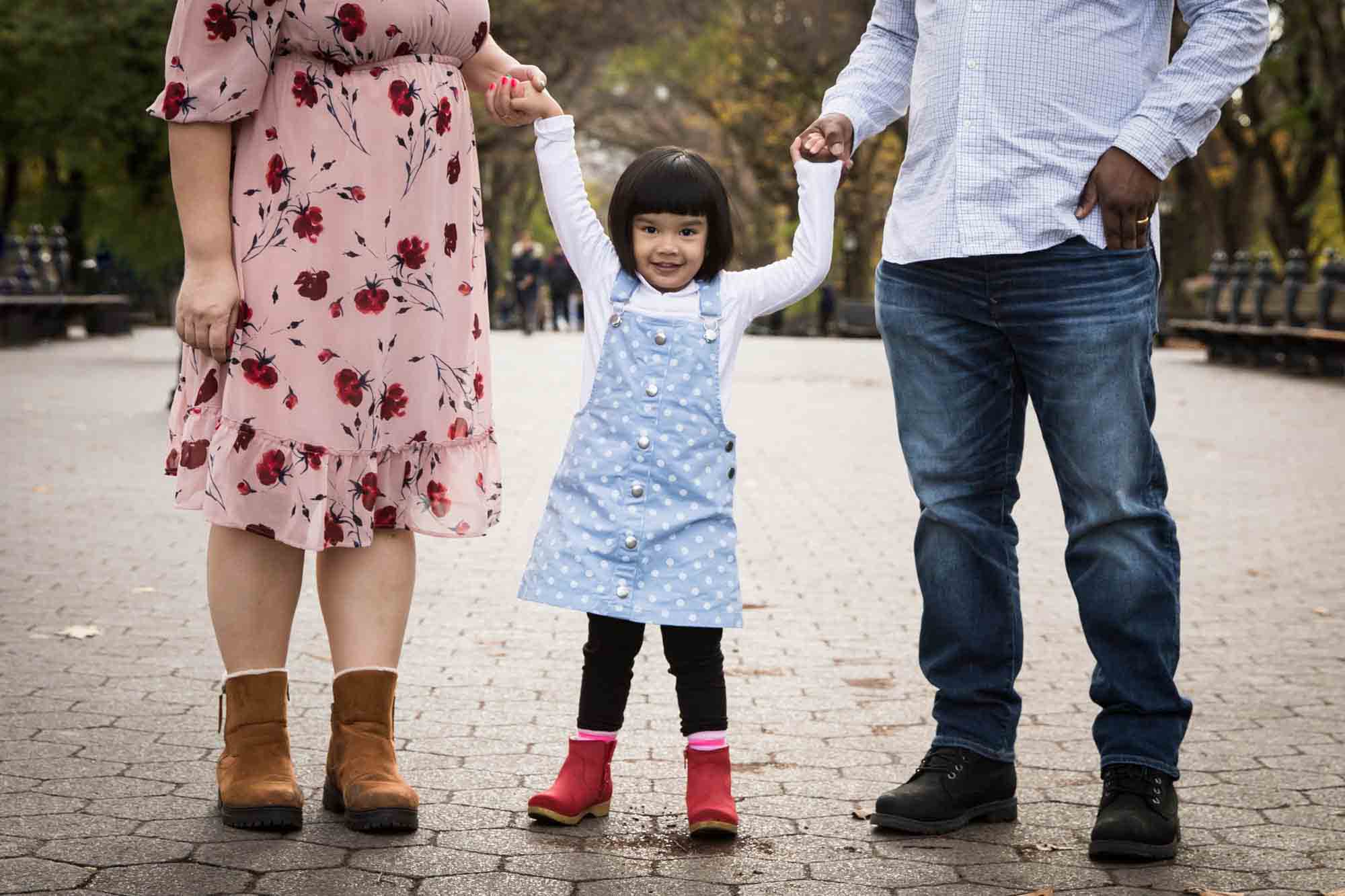 Little girl holding hands with parents in pathway during Central Park family portrait