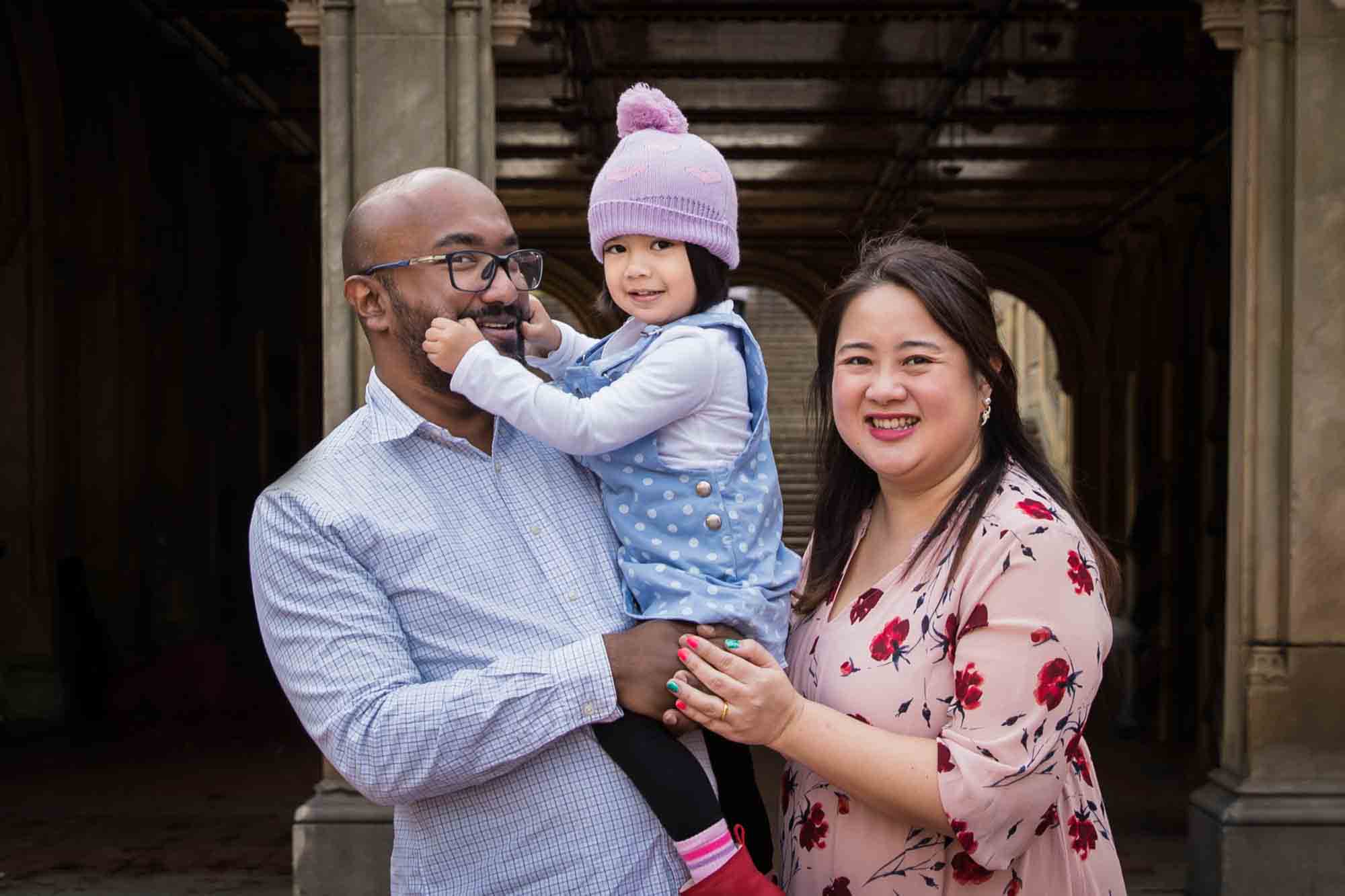 Parents holding little girl who is holding on to father's mustache for an article on Central Park winter portrait tips