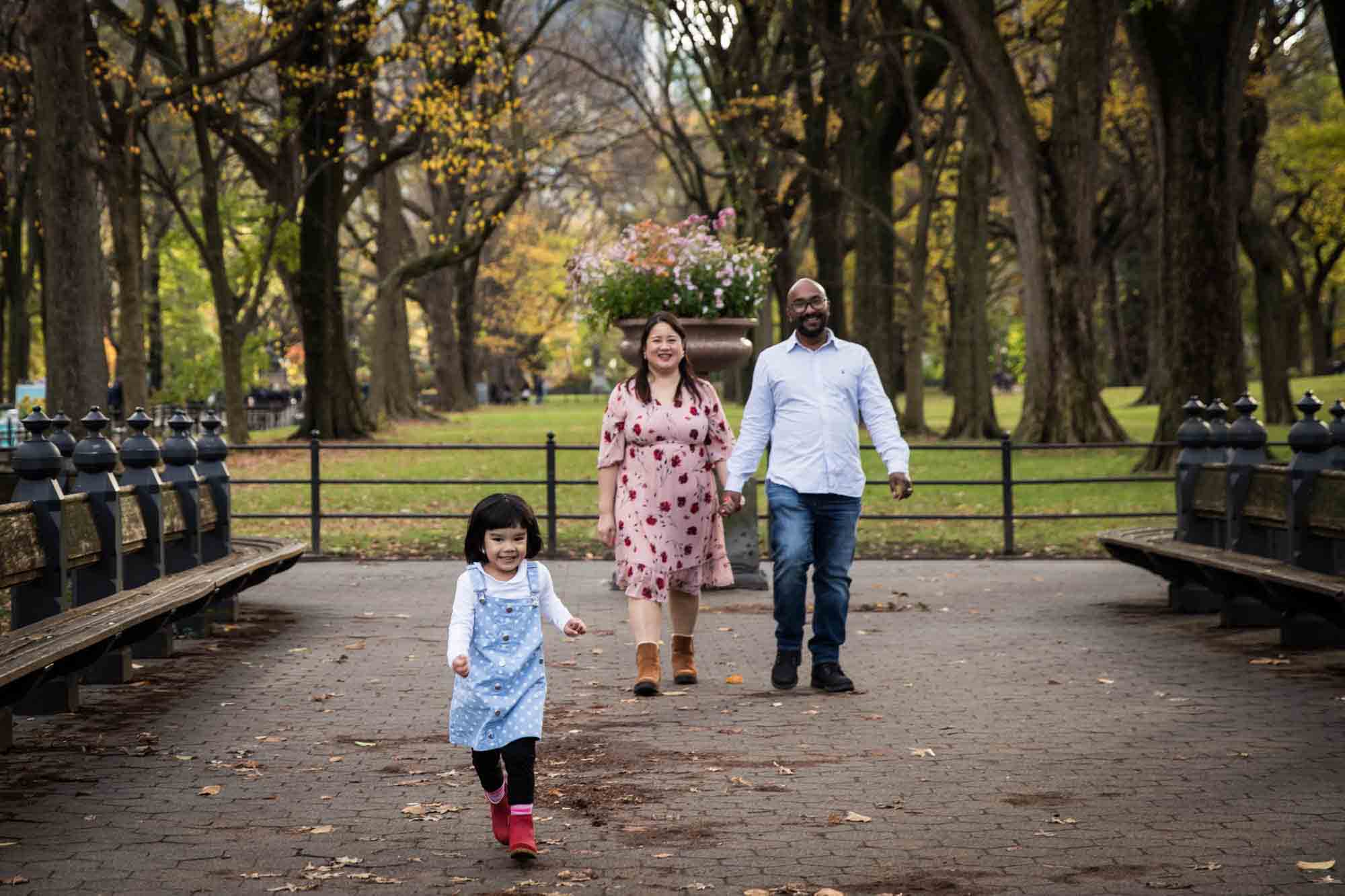 Little girl running down pathway with parents walking behind for an article on Central Park winter portrait tips