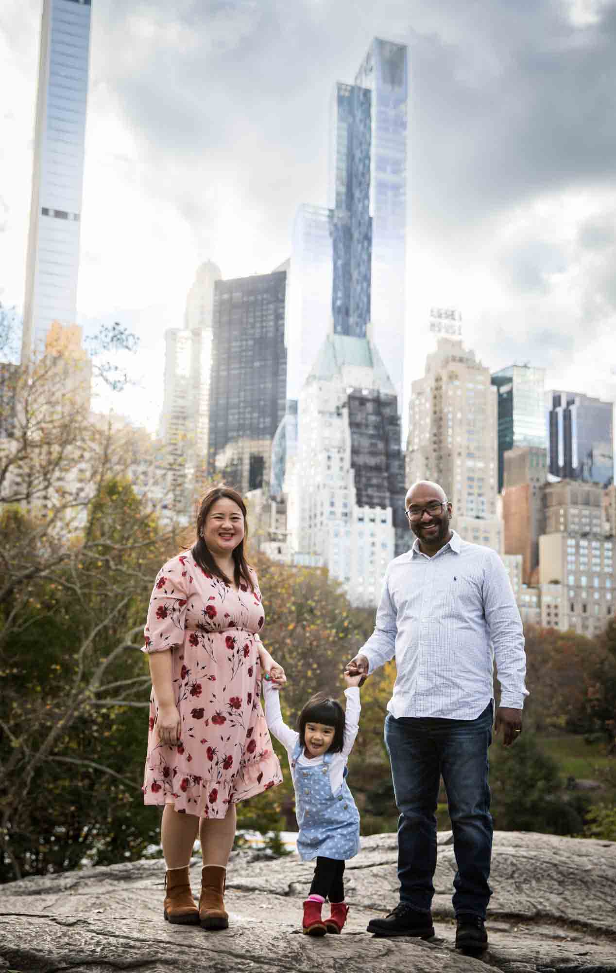 Central Park family portrait of parents on rock holding hands of little girl with NYC skyline in the background