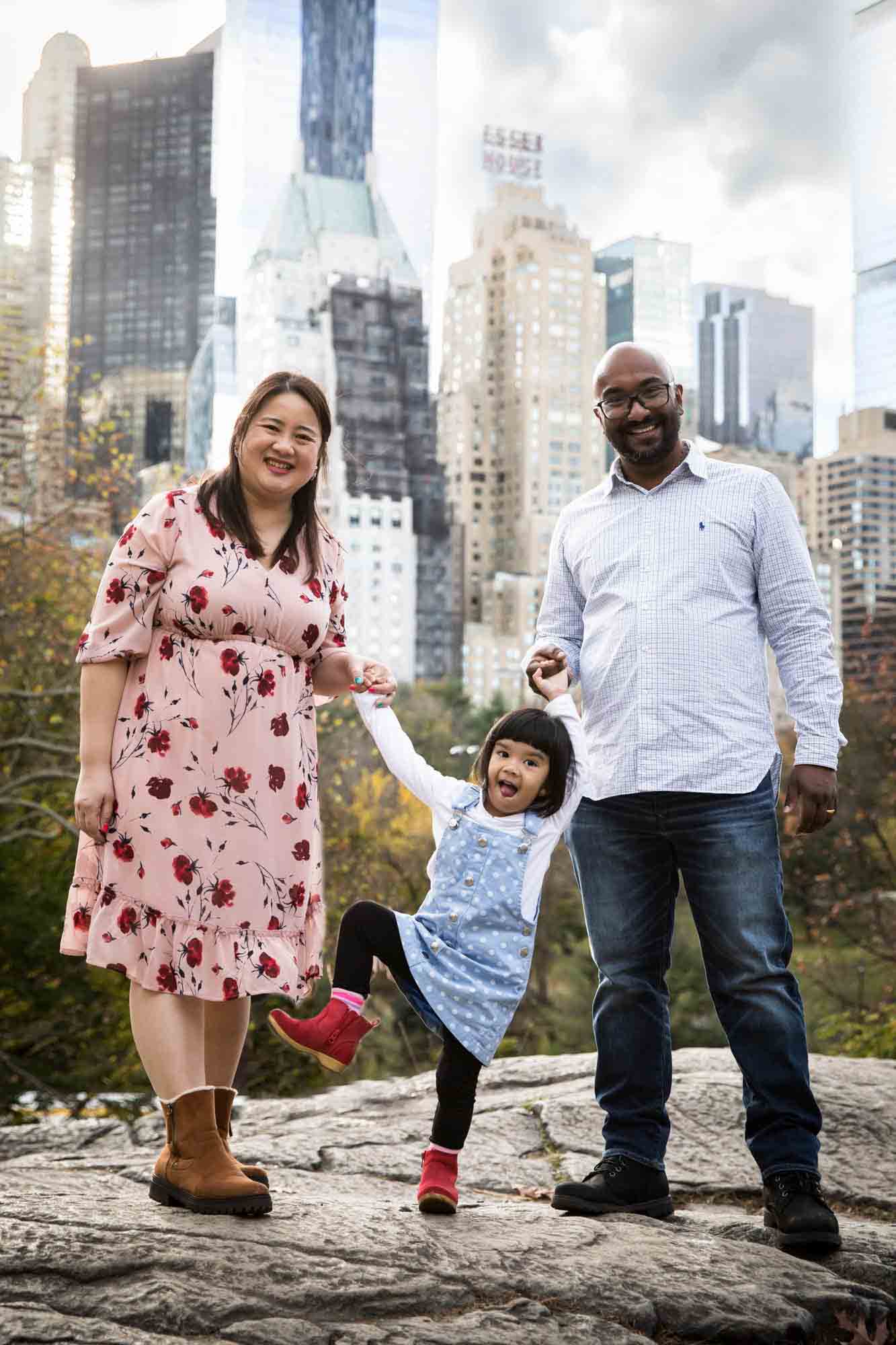 Central Park family portrait of parents on rock holding hands of little girl with NYC skyline in the background