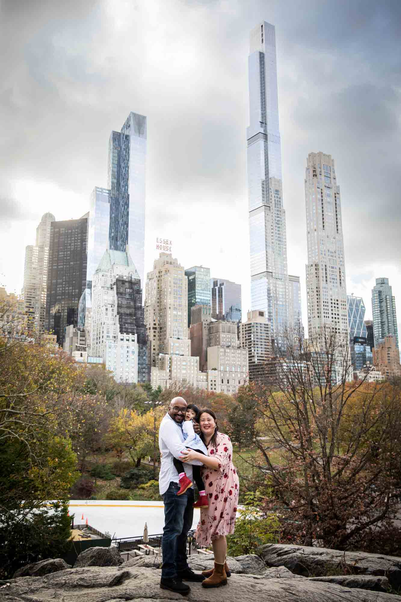 Central Park family portrait of parents on rock holding little girl with NYC skyline in the background