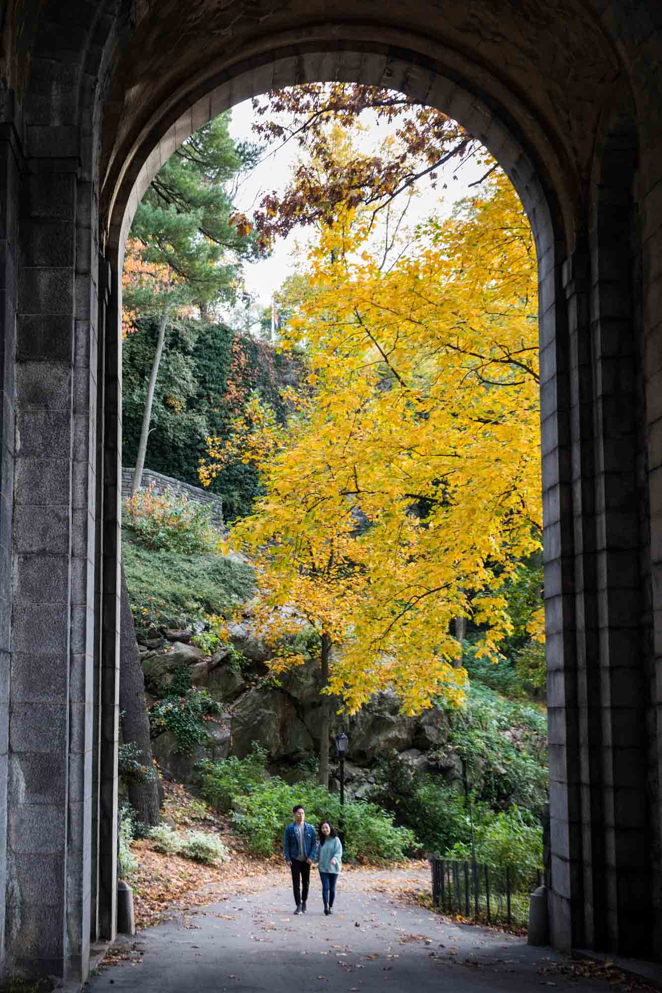 Couple walking under Billings Arcade in front of tree with yellow leaves at a Fort Tryon Park engagement photo shoot