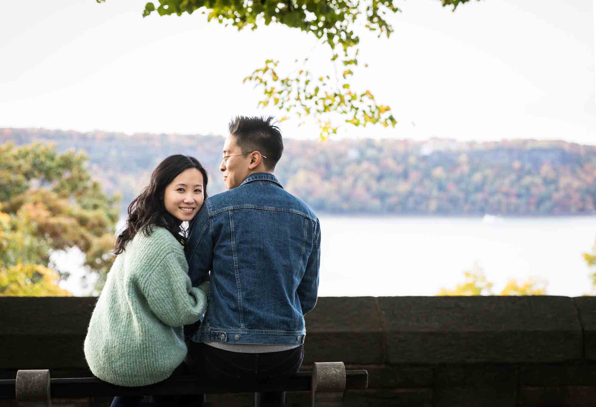 Couple sitting on bench with view of Hudson River at a Fort Tryon Park engagement photo shoot