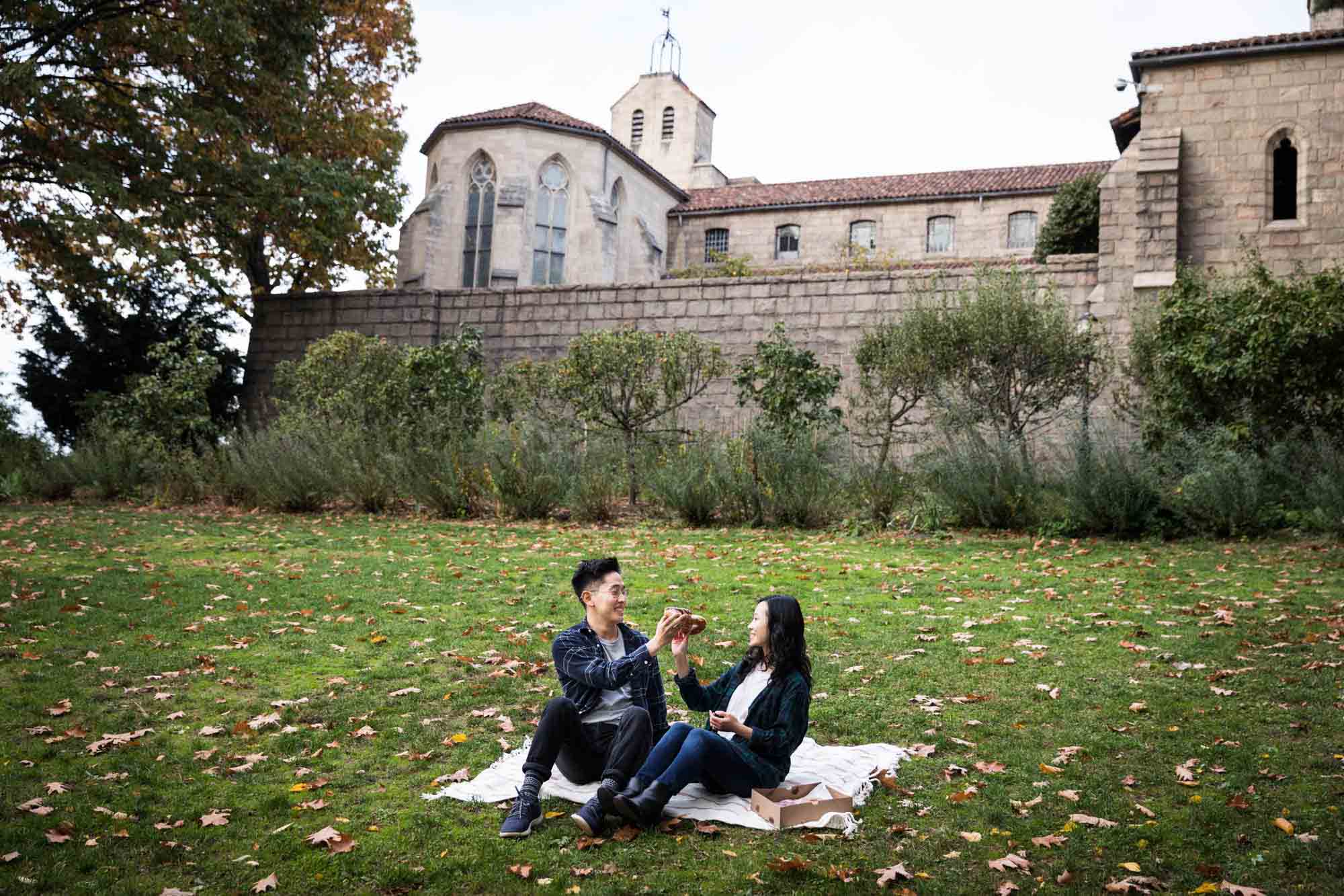 Fort Tryon Park engagement photos of couple eating doughnuts on picnic blanket in front of Cloisters Museum