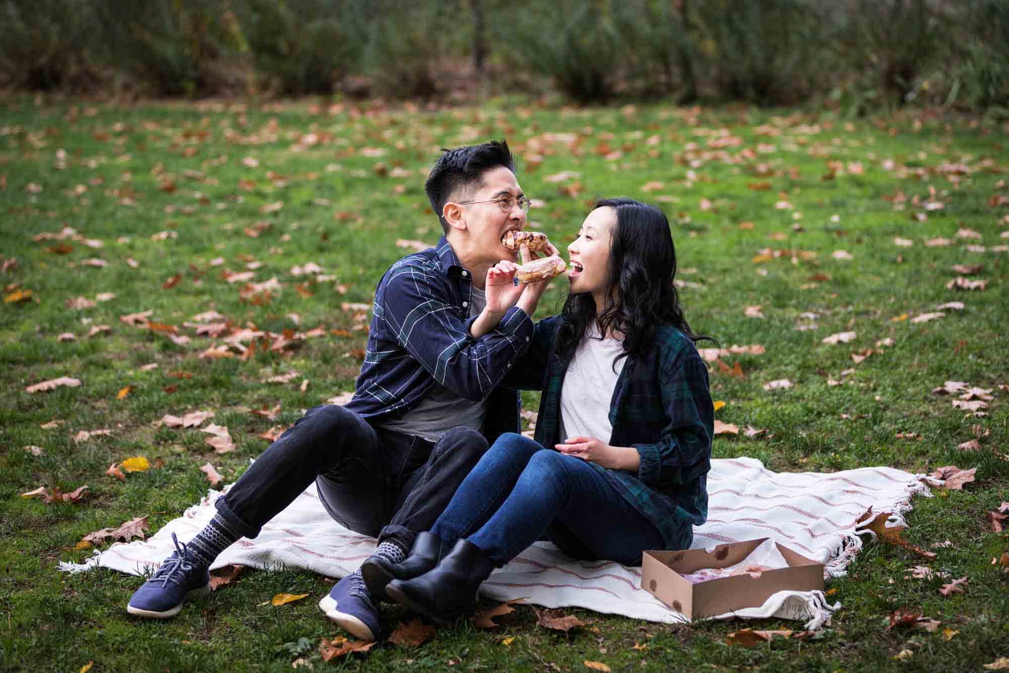 Fort Tryon Park engagement photo of couple eating doughnuts on picnic blanket with intertwined arms