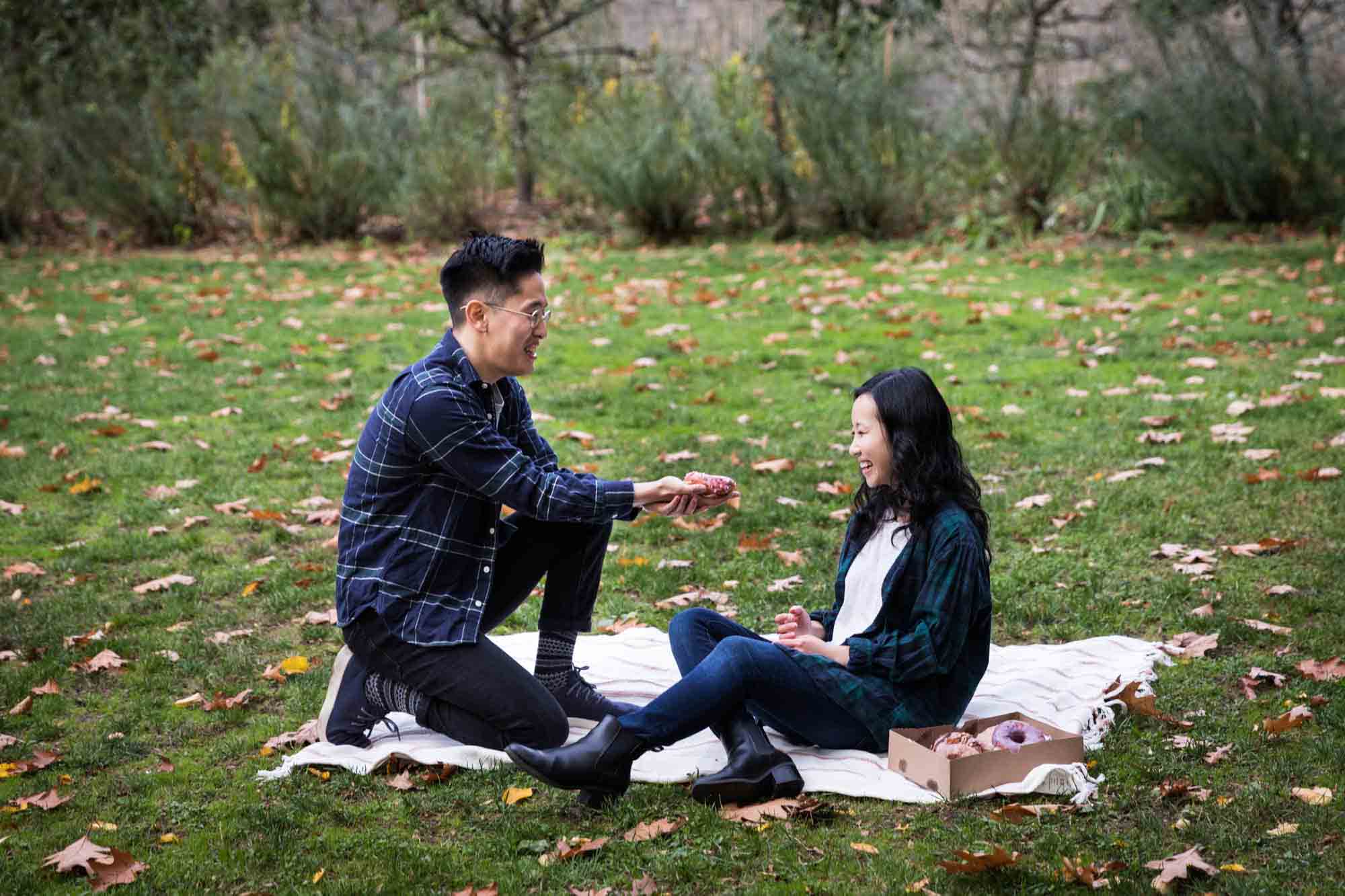 Fort Tryon Park engagement photo of man proposing to woman on picnic blanket with doughnut