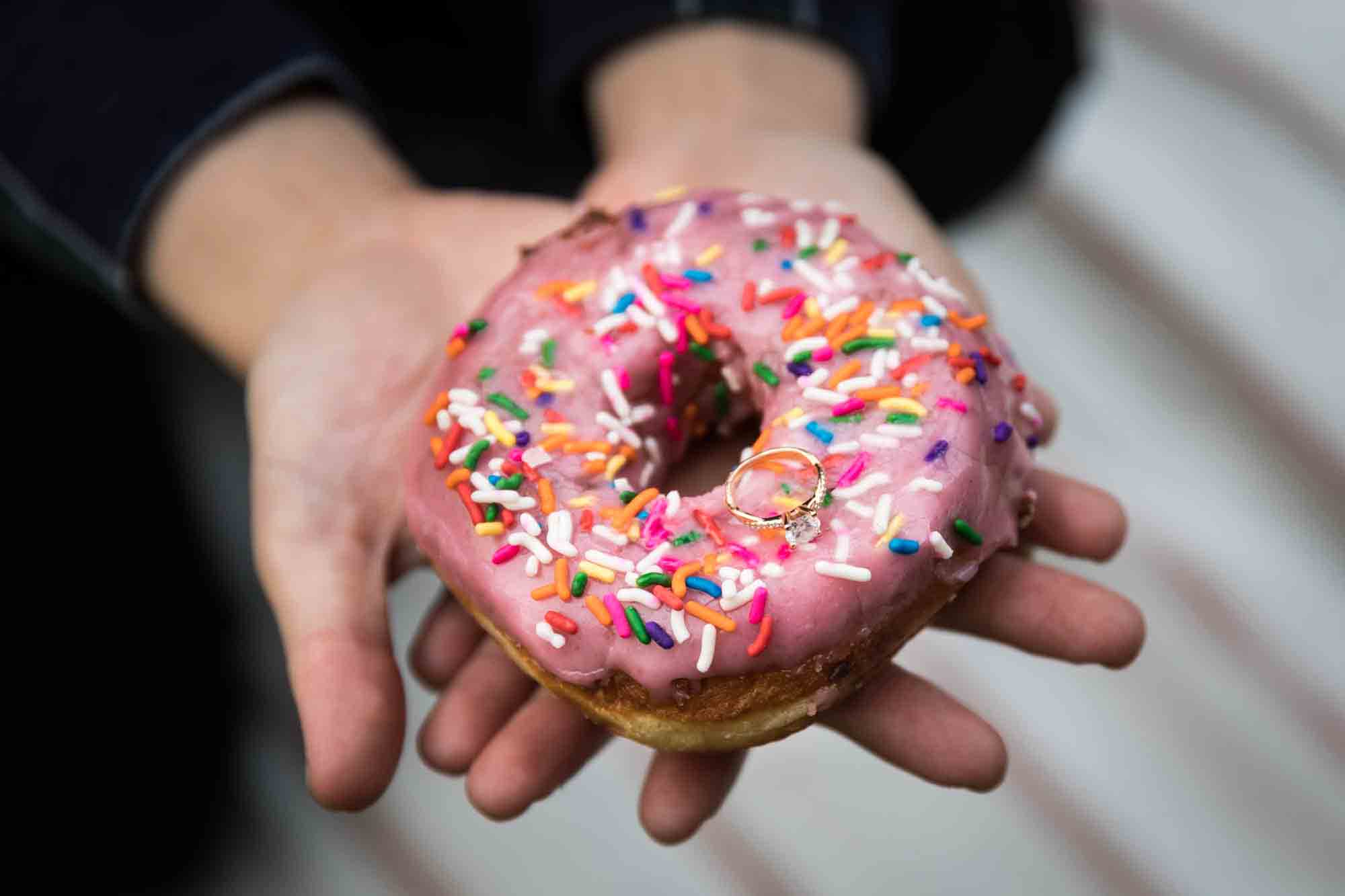 Man's hands offering engagement ring on top of doughnut covered in pink glaze and colorful sprinkles
