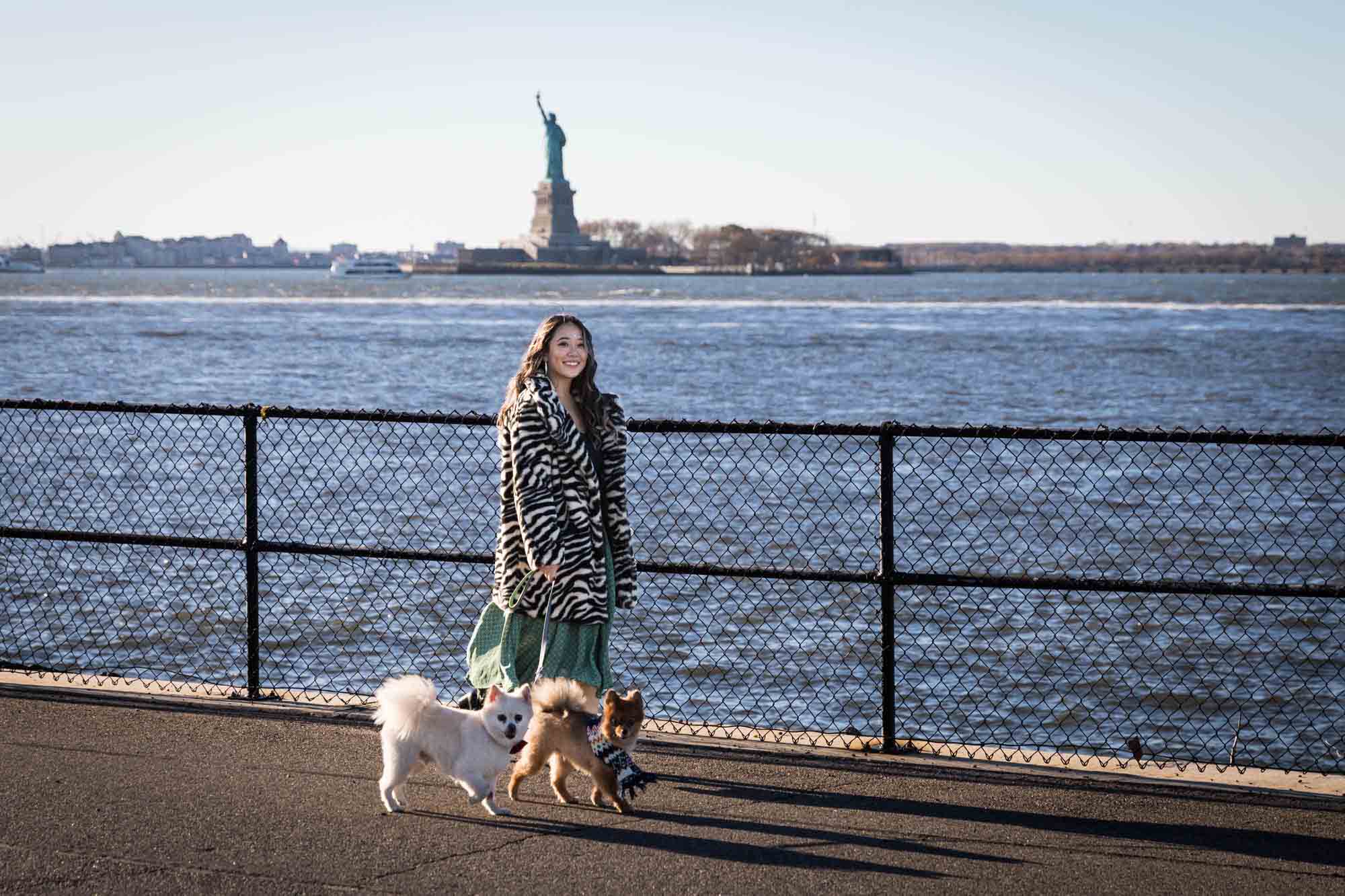 Pet portraits on Governors Island of woman walking in front of fence with two Pomeranian dogs and Statue of Liberty in background