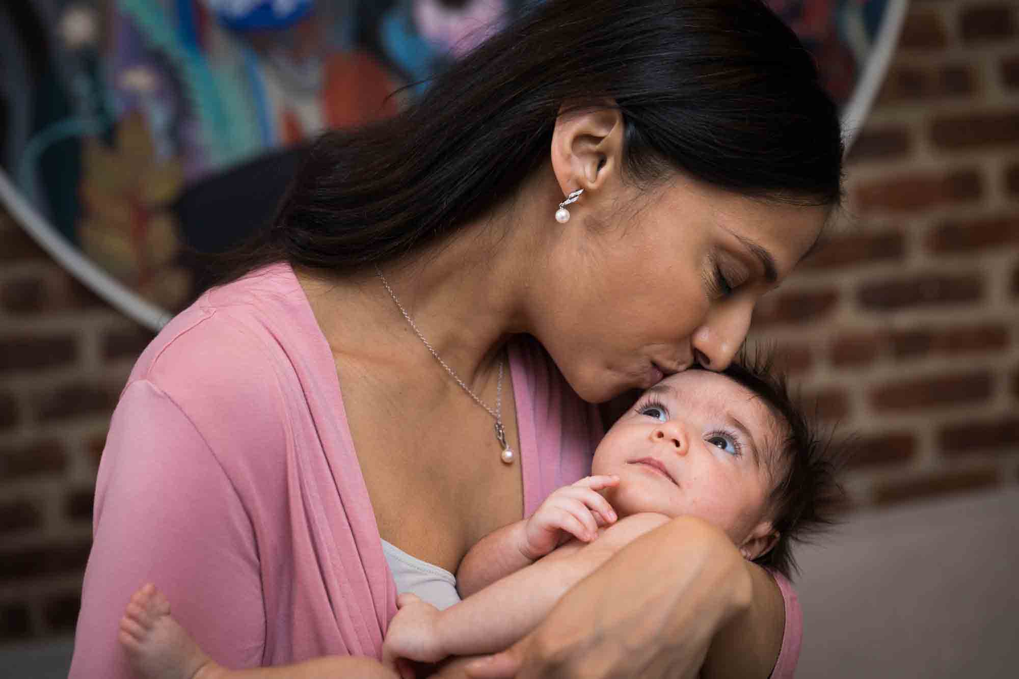 Mother kissing newborn baby on forehead in front of brick wall