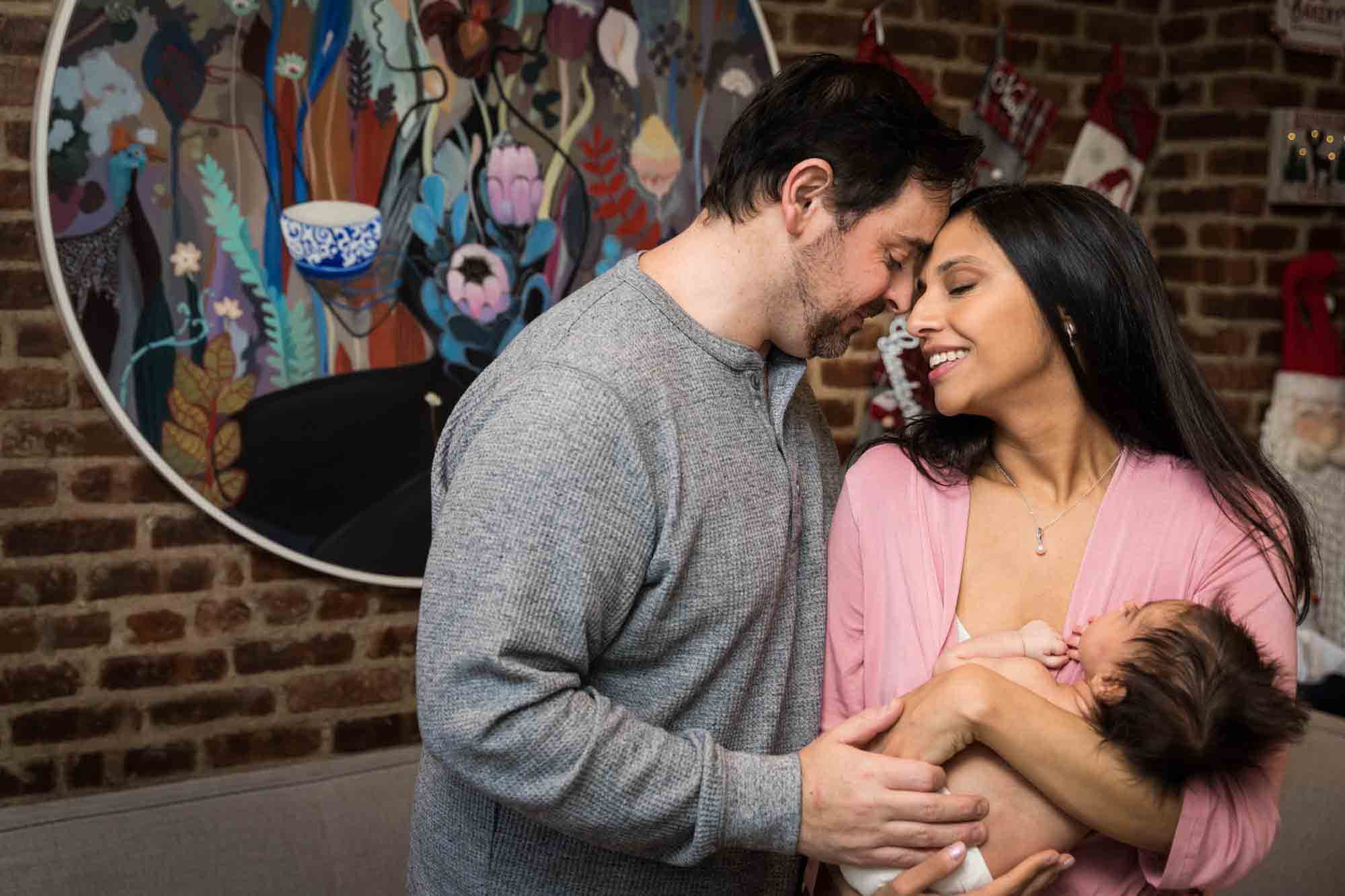 Parents touching foreheads while holding newborn baby in front of brick wall