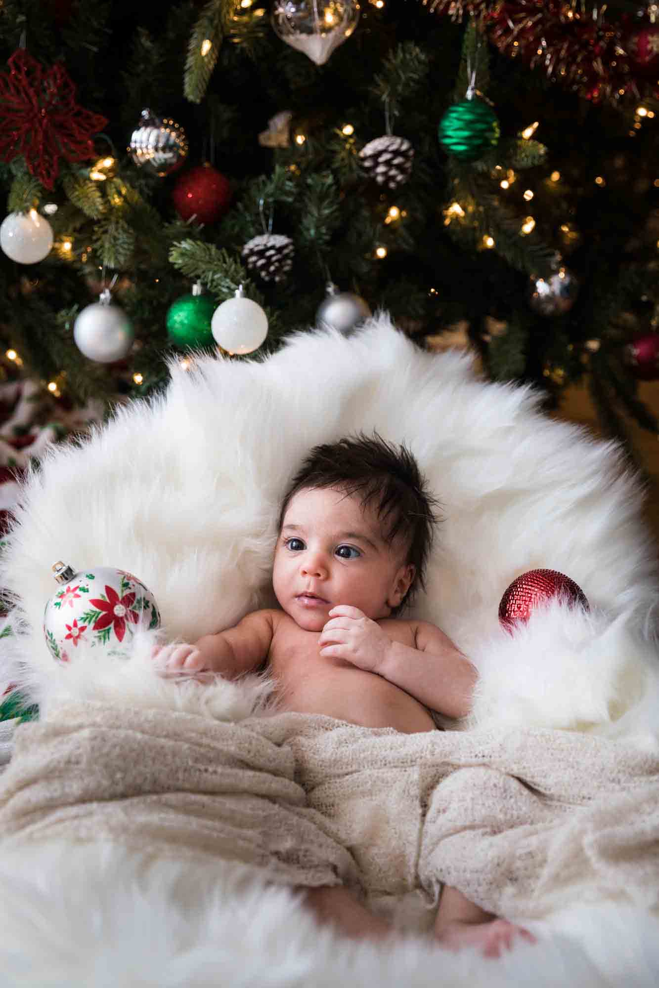 Newborn baby on white rug looking at Christmas ornaments in front of tree