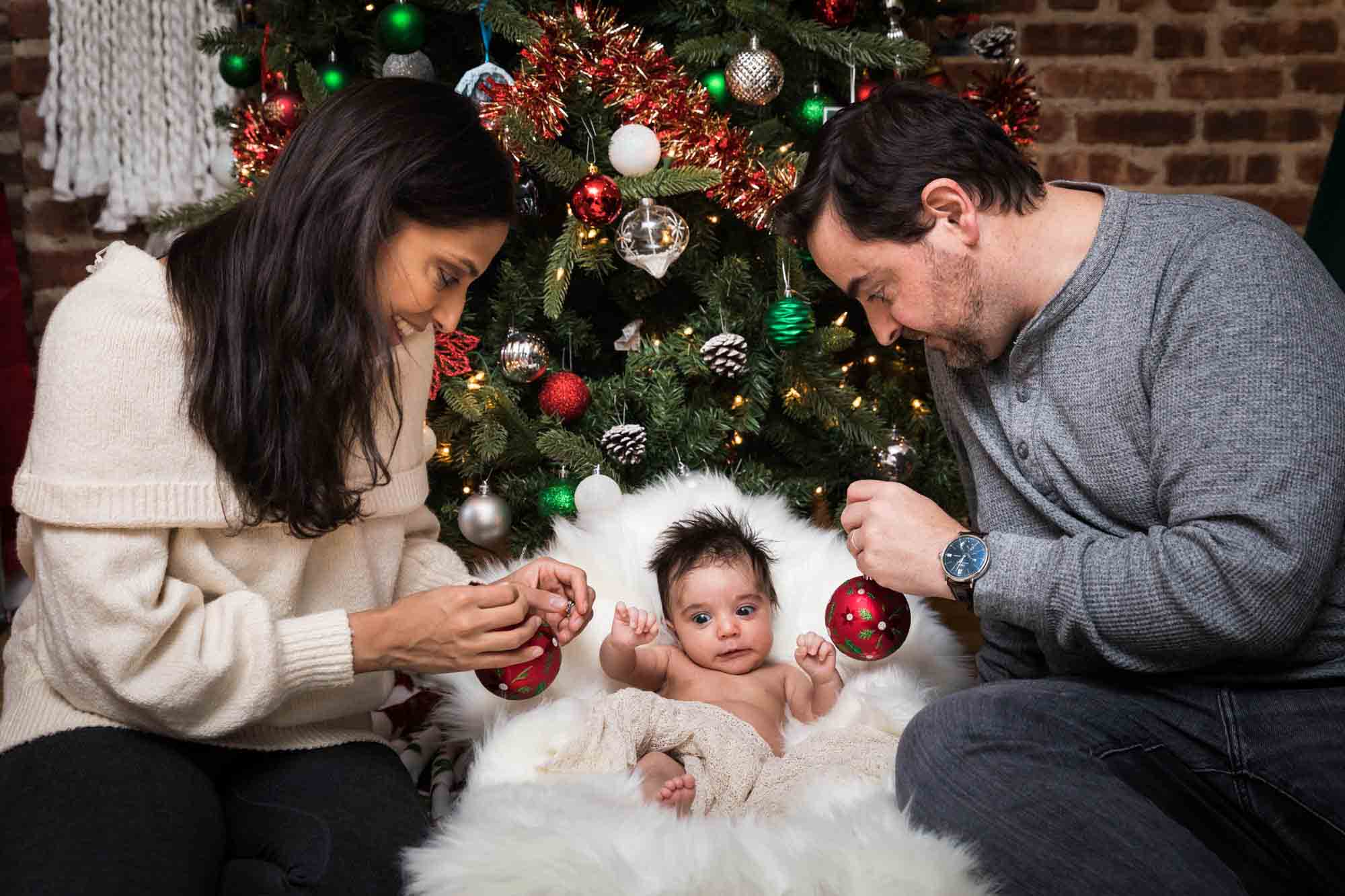 Parents holding Christmas ornaments in front of newborn baby during a Christmas-themed newborn photo shoot in NYC