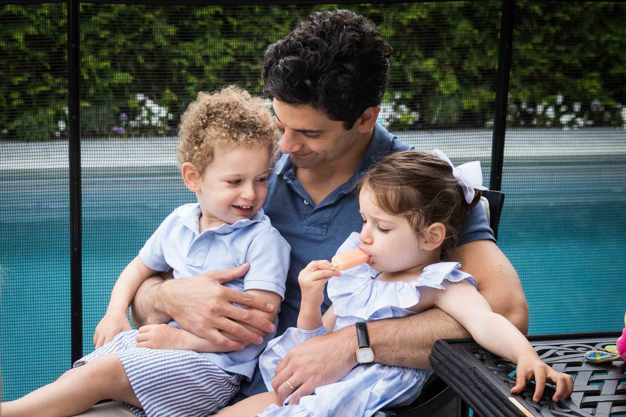 Father holding little girl and boy on his lap in front of pool