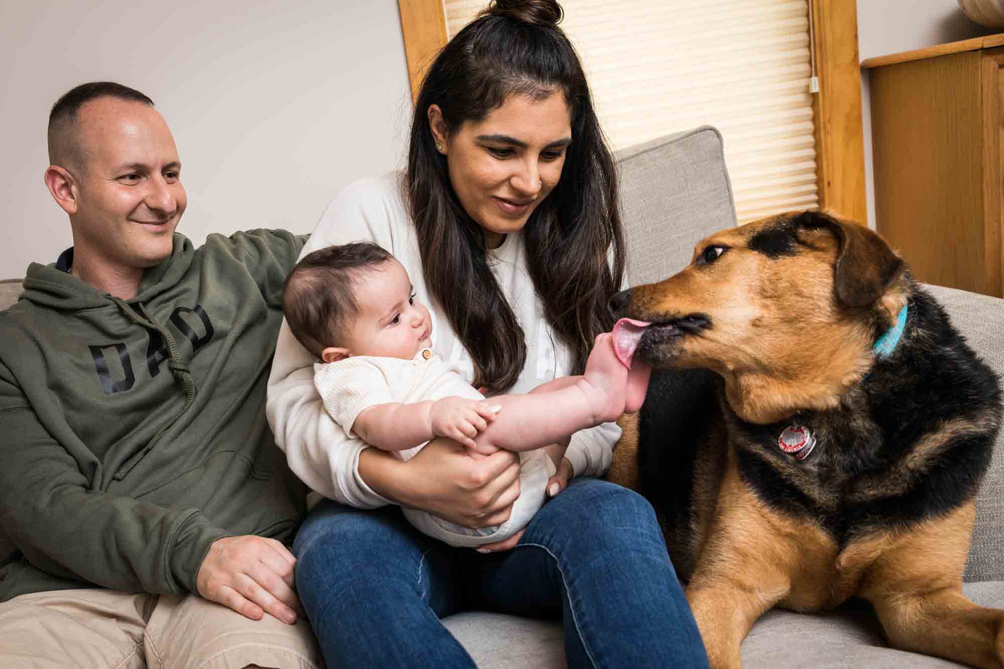 Mother holding baby on couch while dog licks baby's feet