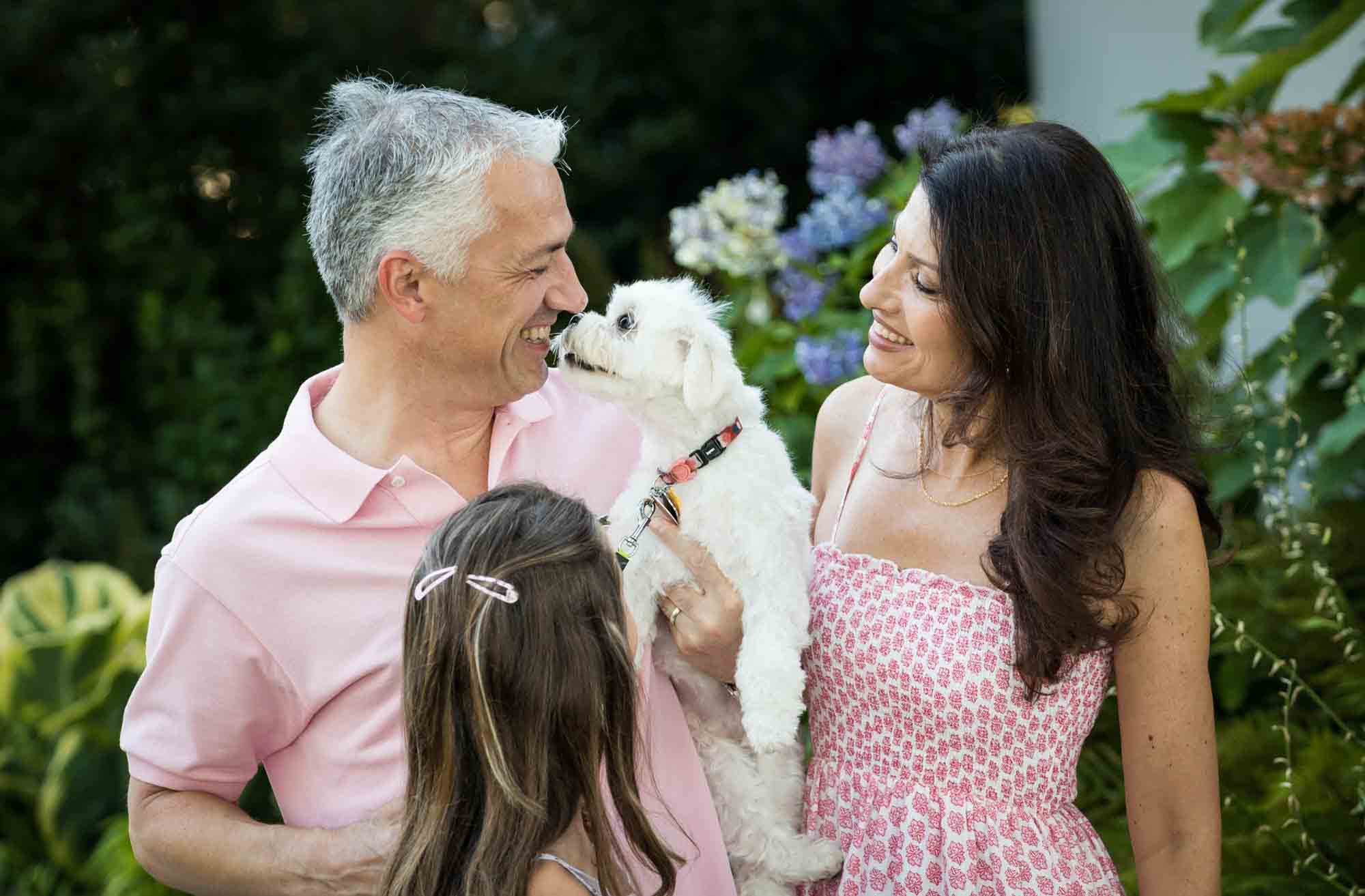 Family watching as white dog licks man on the face 