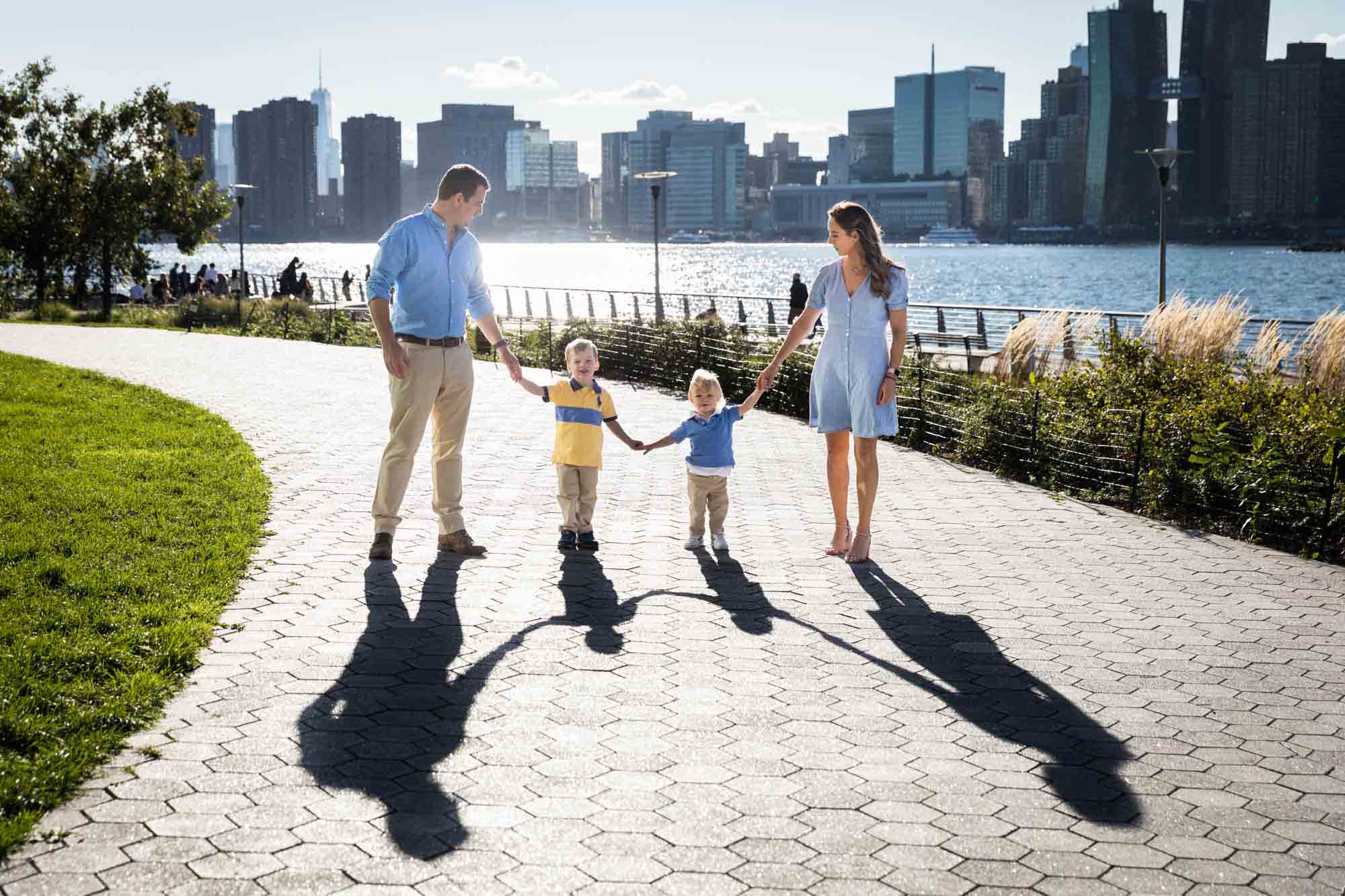 Parents holding hands of two young boys and walking with shadow in Gantry Plaza State Park