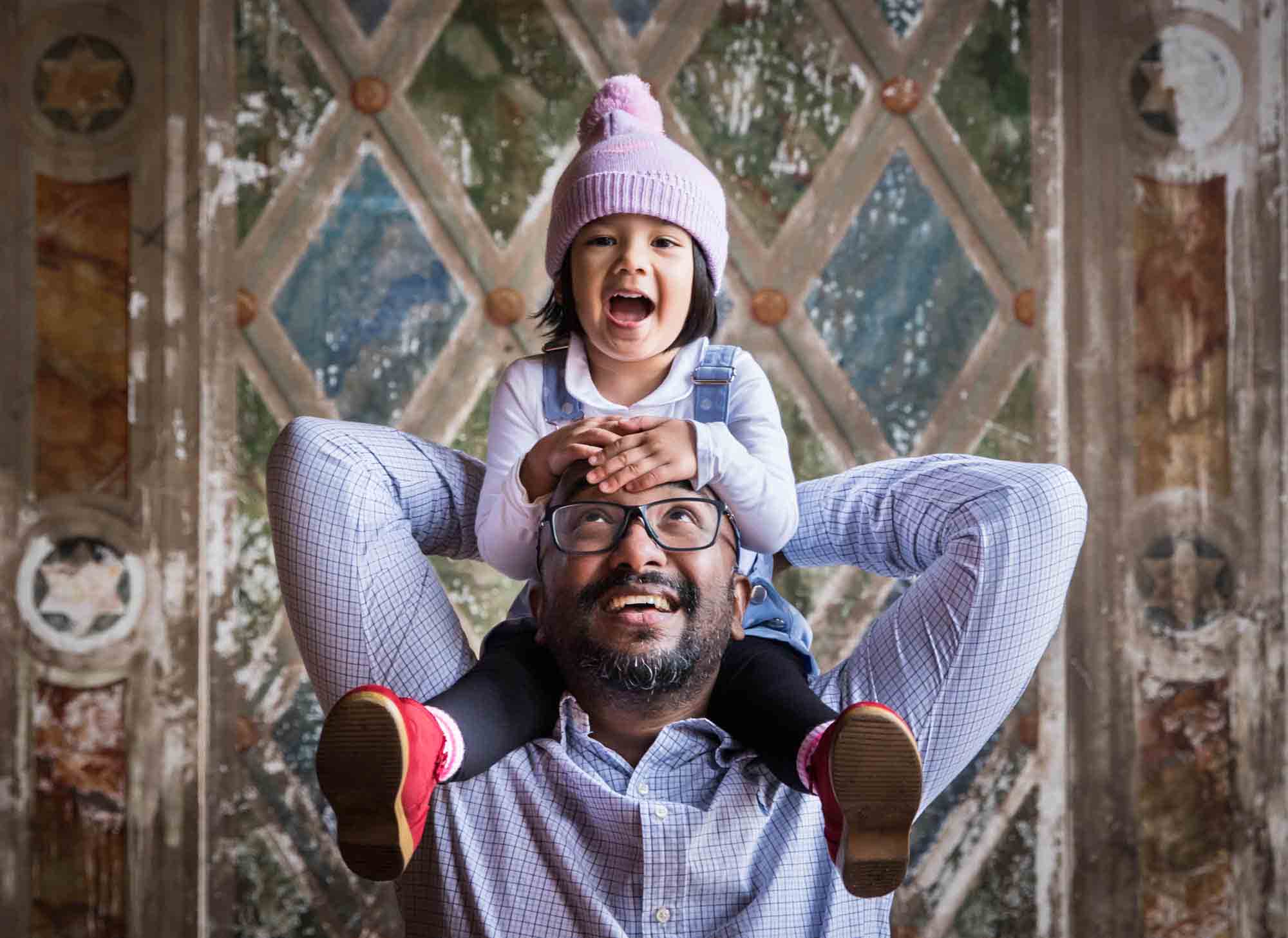 Father holding little girl wearing pink knitted cap on his shoulders 