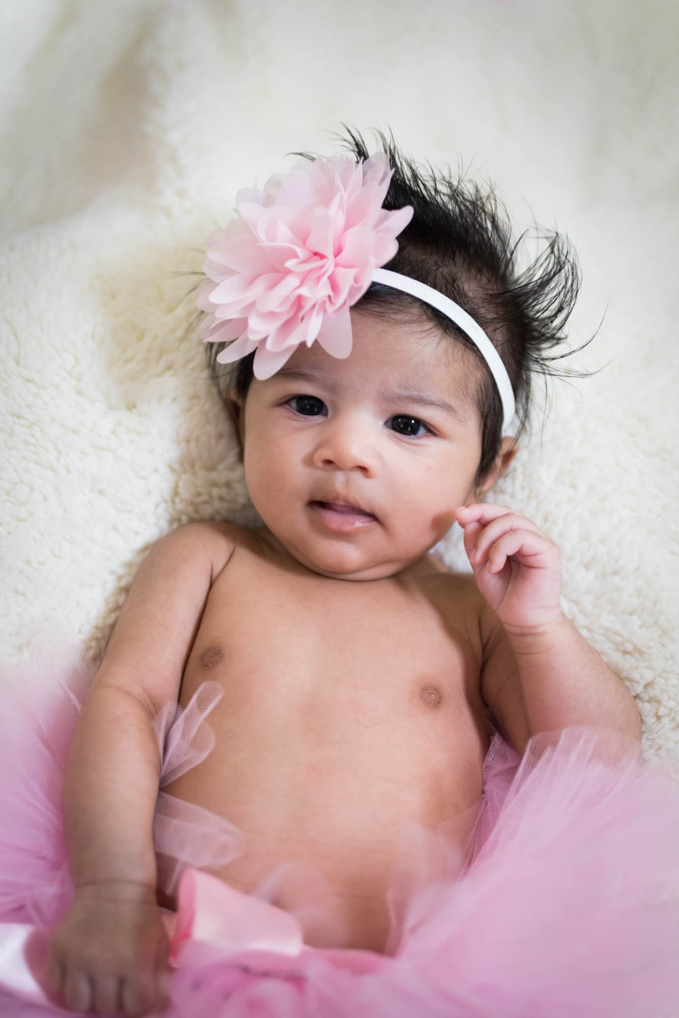 Little newborn girl lying on white fur rug wearing pink flower in her hair for an article advertising family portrait gift certificates