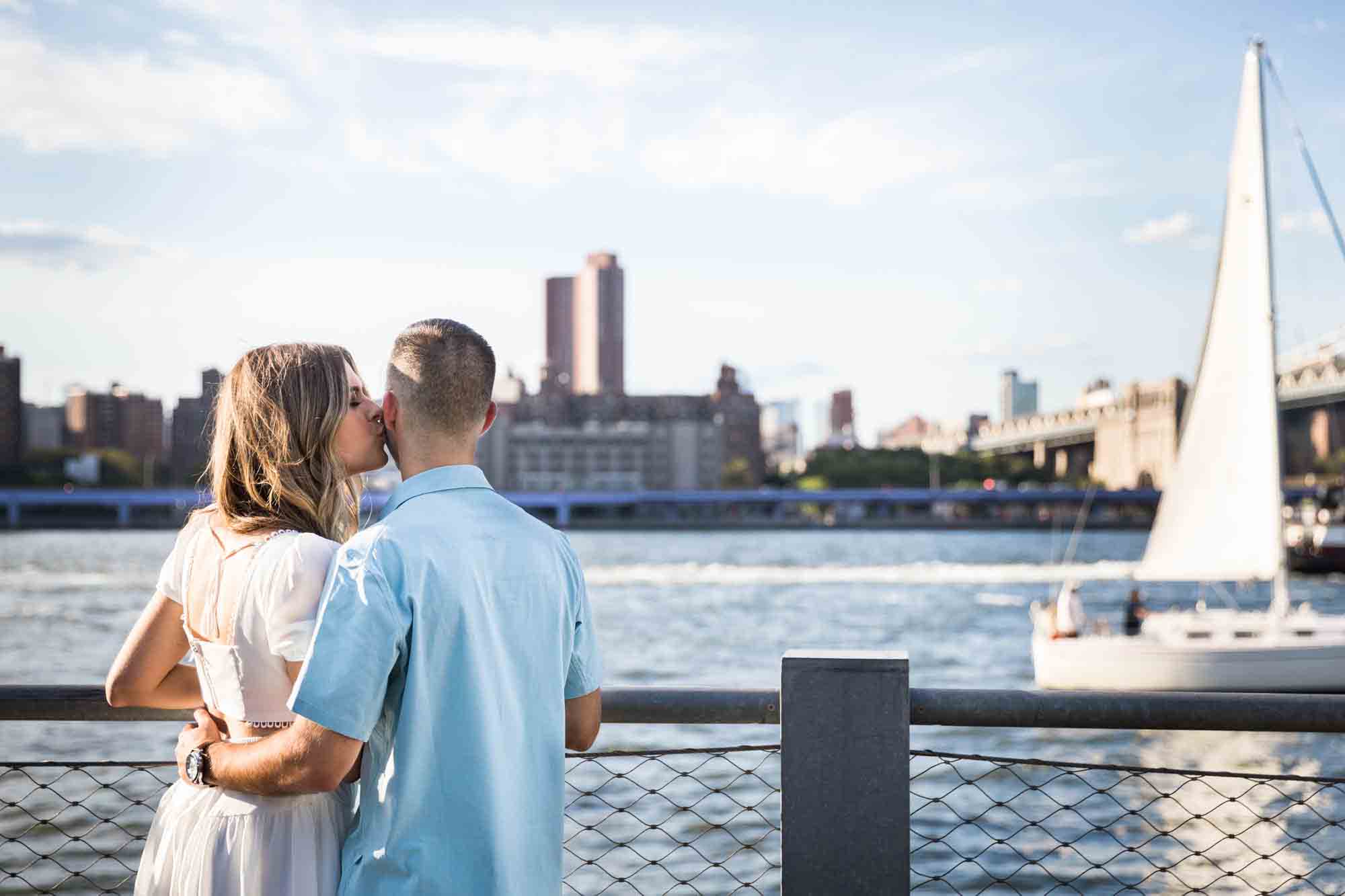 Woman kissing man on the cheek in front of East River waterfront during a Brooklyn Bridge Park surprise proposal