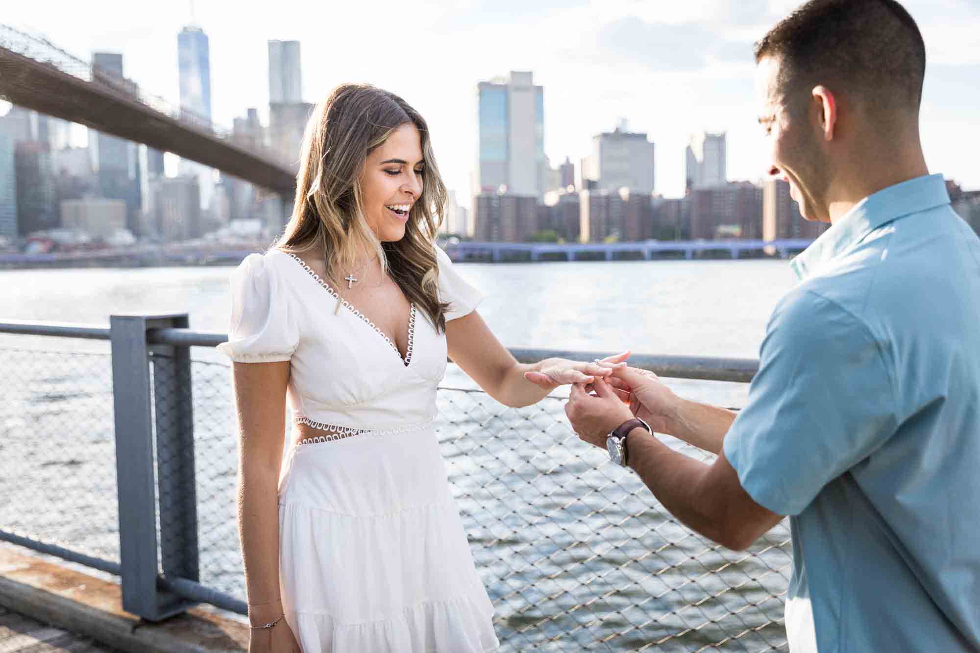 Man putting ring on woman's hand in front of East River during a surprise proposal in Brooklyn Bridge Park