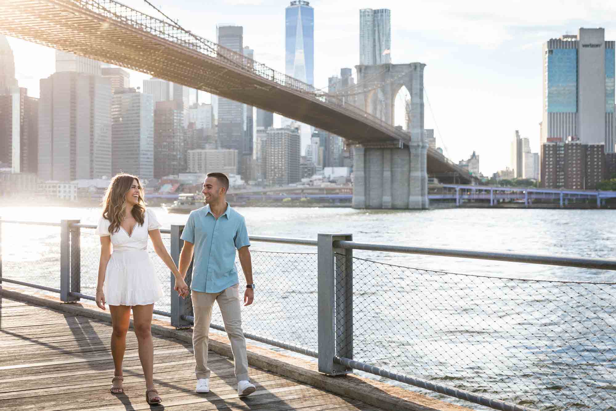 Couple holding hands and walking along boardwalk in front of Brooklyn Bridge during a surprise proposal in Brooklyn Bridge Park