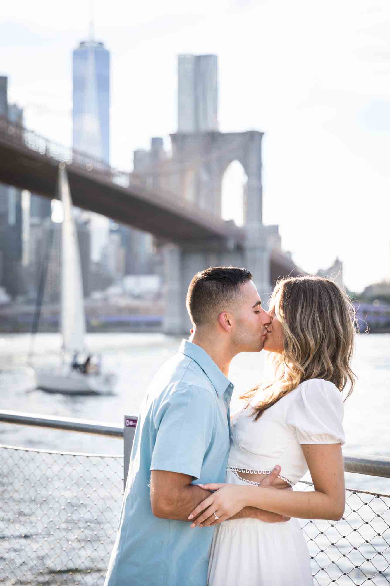 Couple kissing in front of East River waterfront and sailboat during a surprise proposal in Brooklyn Bridge Park