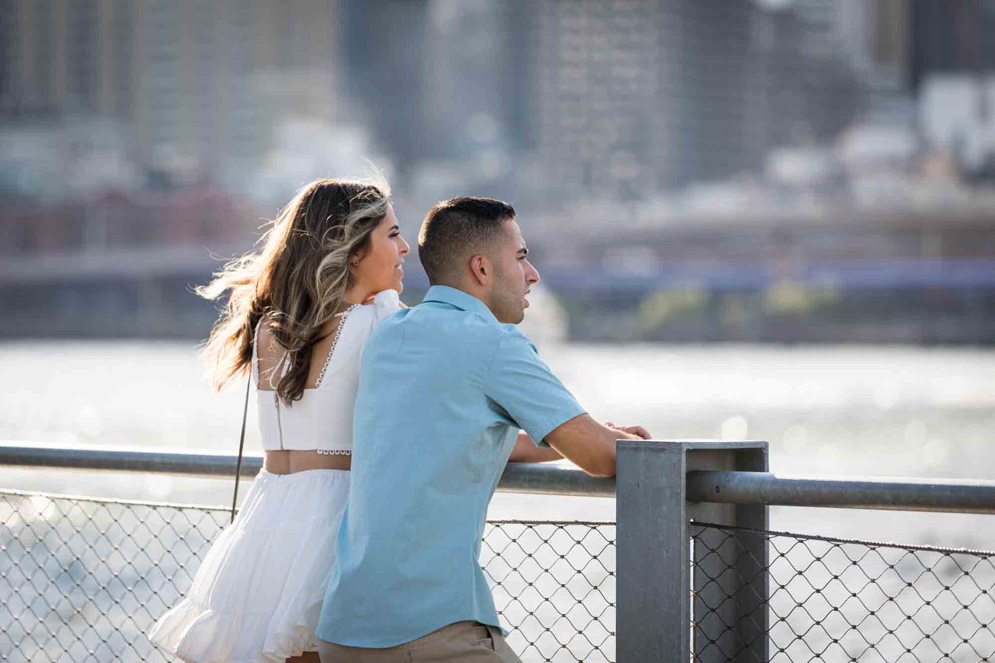Couple leaning on railing looking out at waterfront during a Brooklyn Bridge Park surprise proposal