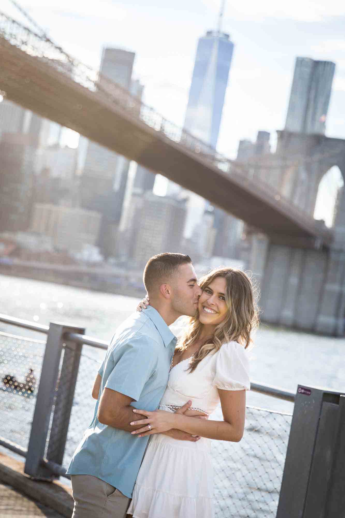 Man kissing woman on the cheek in front of Brooklyn Bridge during a surprise proposal in Brooklyn Bridge Park