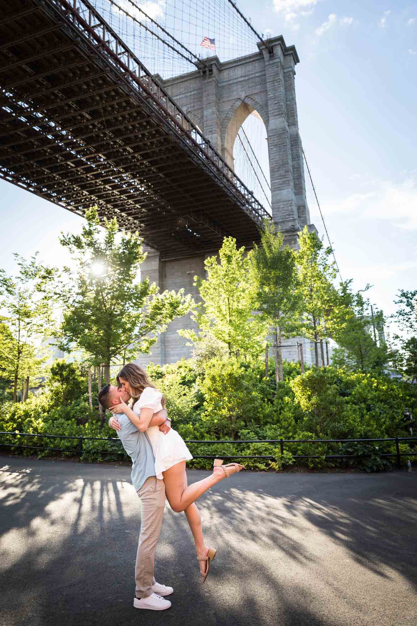 Man lifting woman in the air in front of Brooklyn Bridge during a surprise proposal in Brooklyn Bridge Park