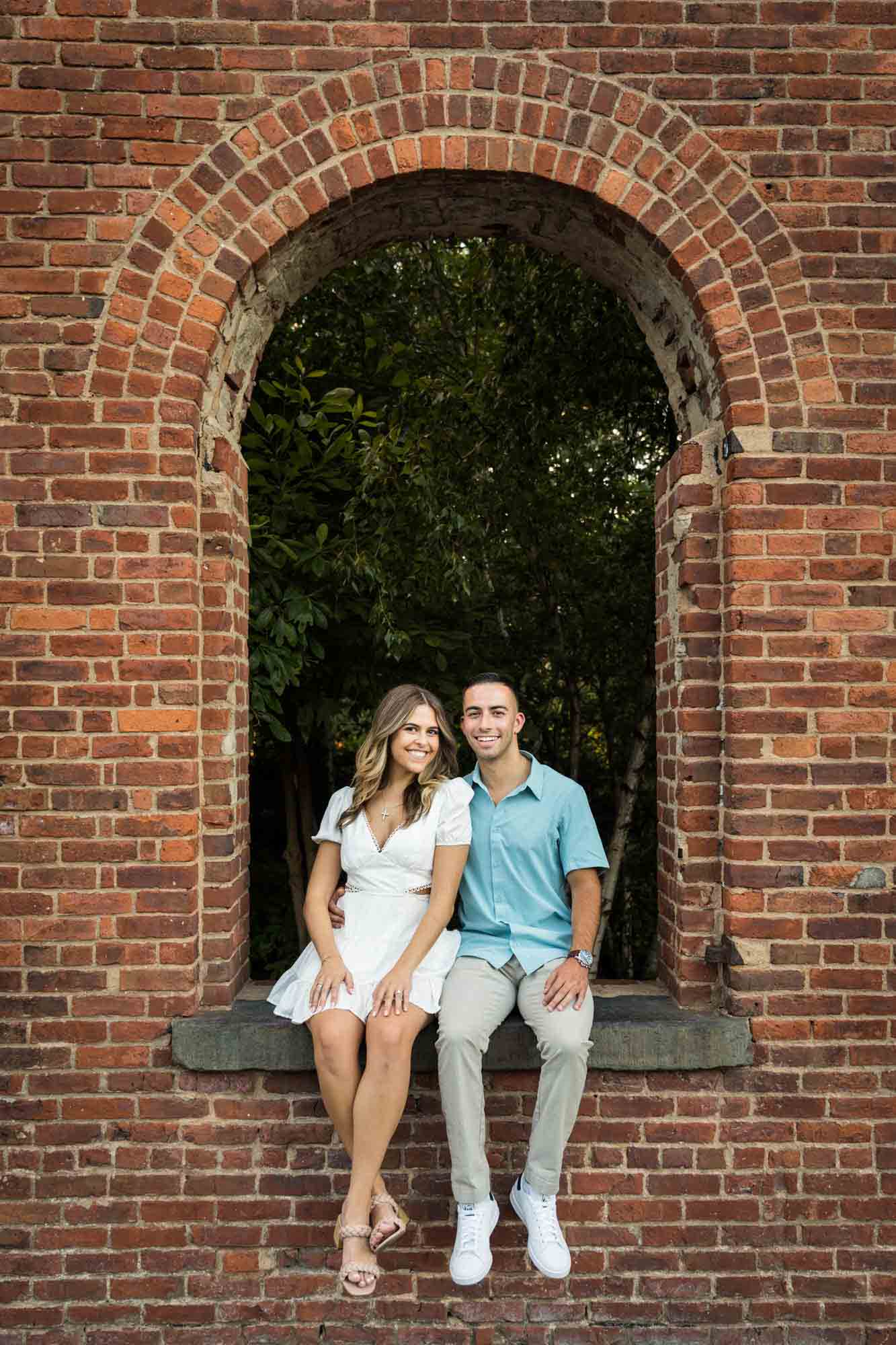 Couple sitting inside window of brick wall during a surprise proposal in Brooklyn Bridge Park