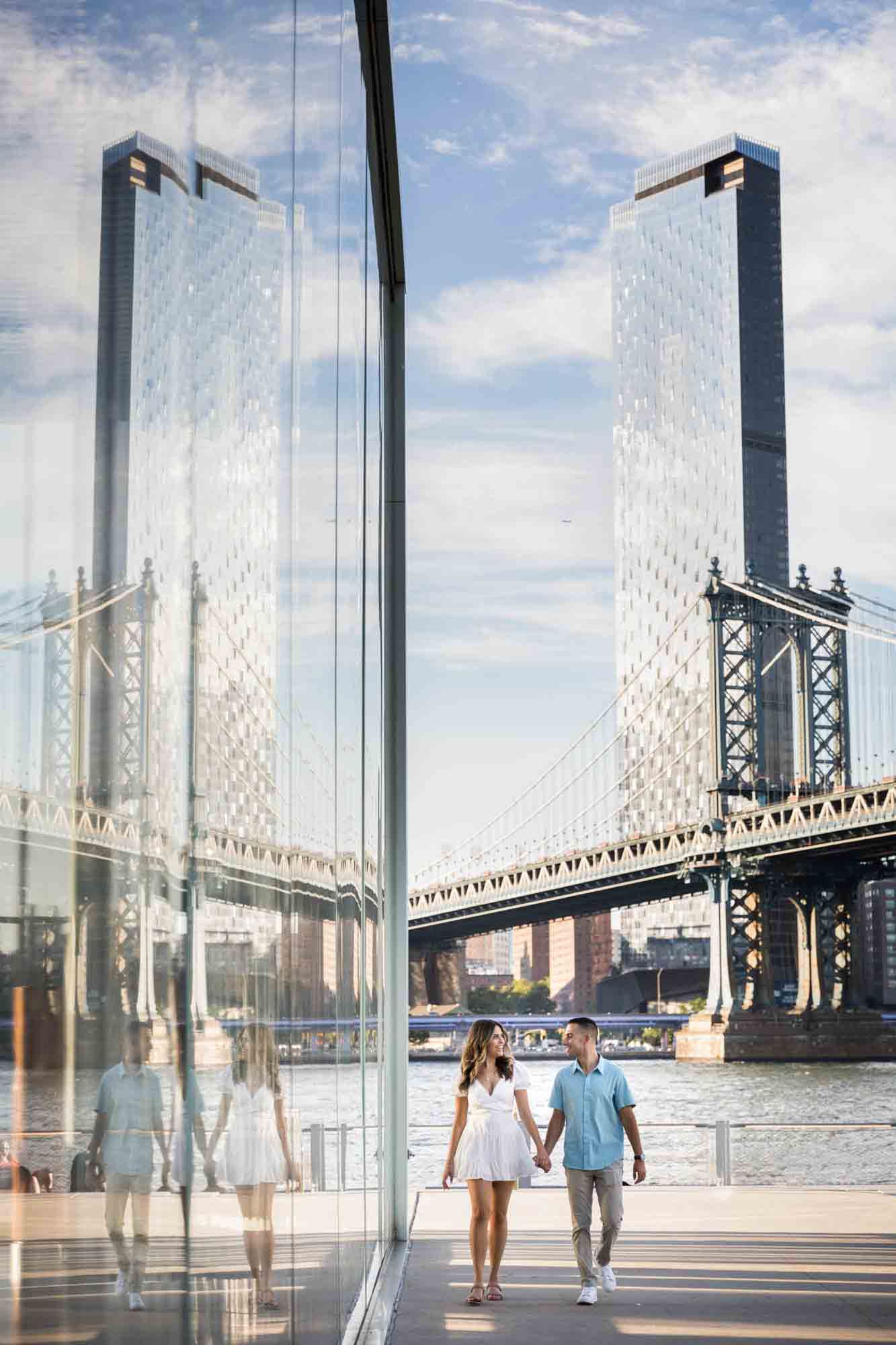Couple reflected in window as they walk along glass wall during a surprise proposal in Brooklyn Bridge Park