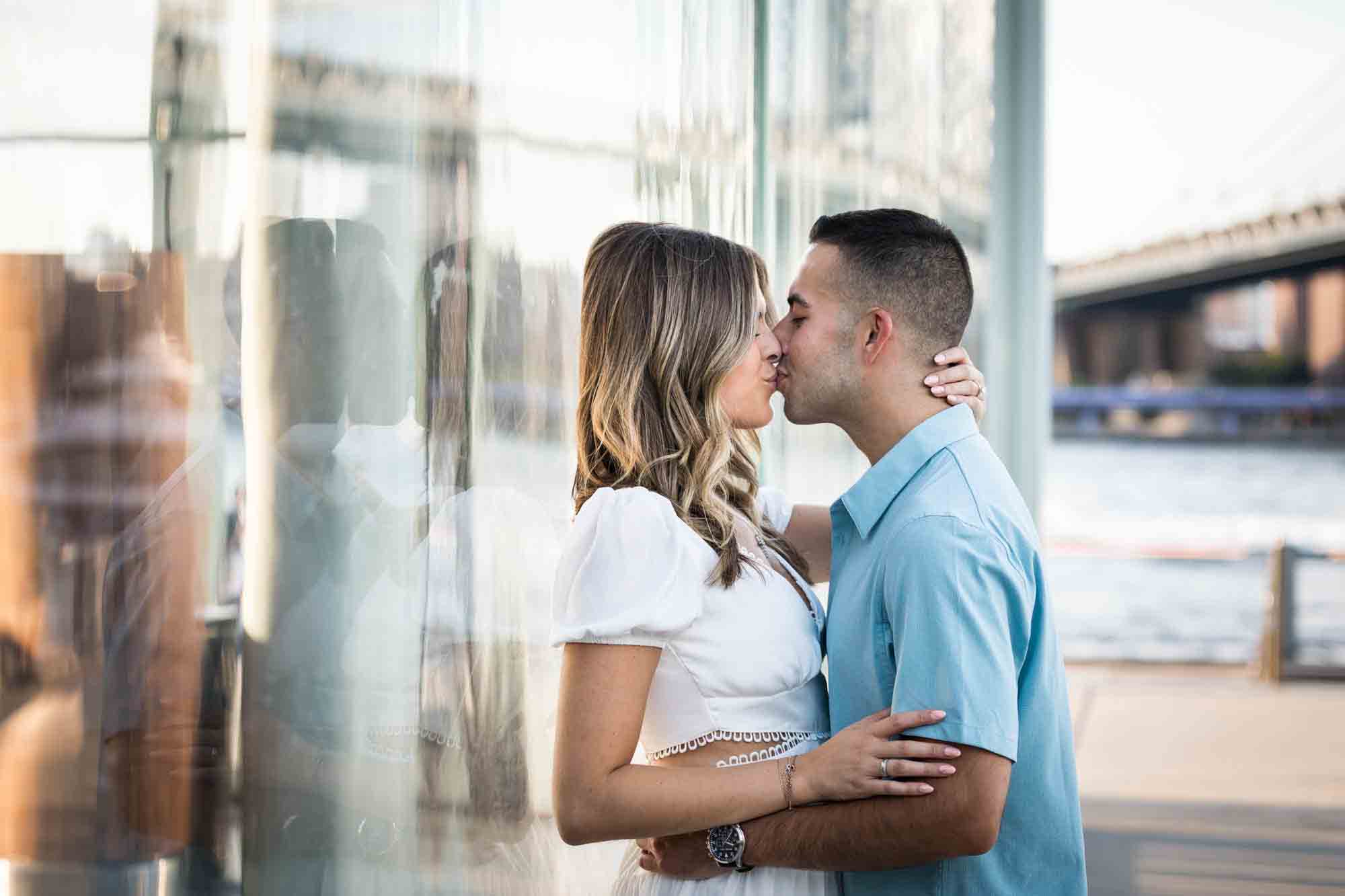 Couple kissing against glass wall of Jane's Carousel during a surprise proposal in Brooklyn Bridge Park