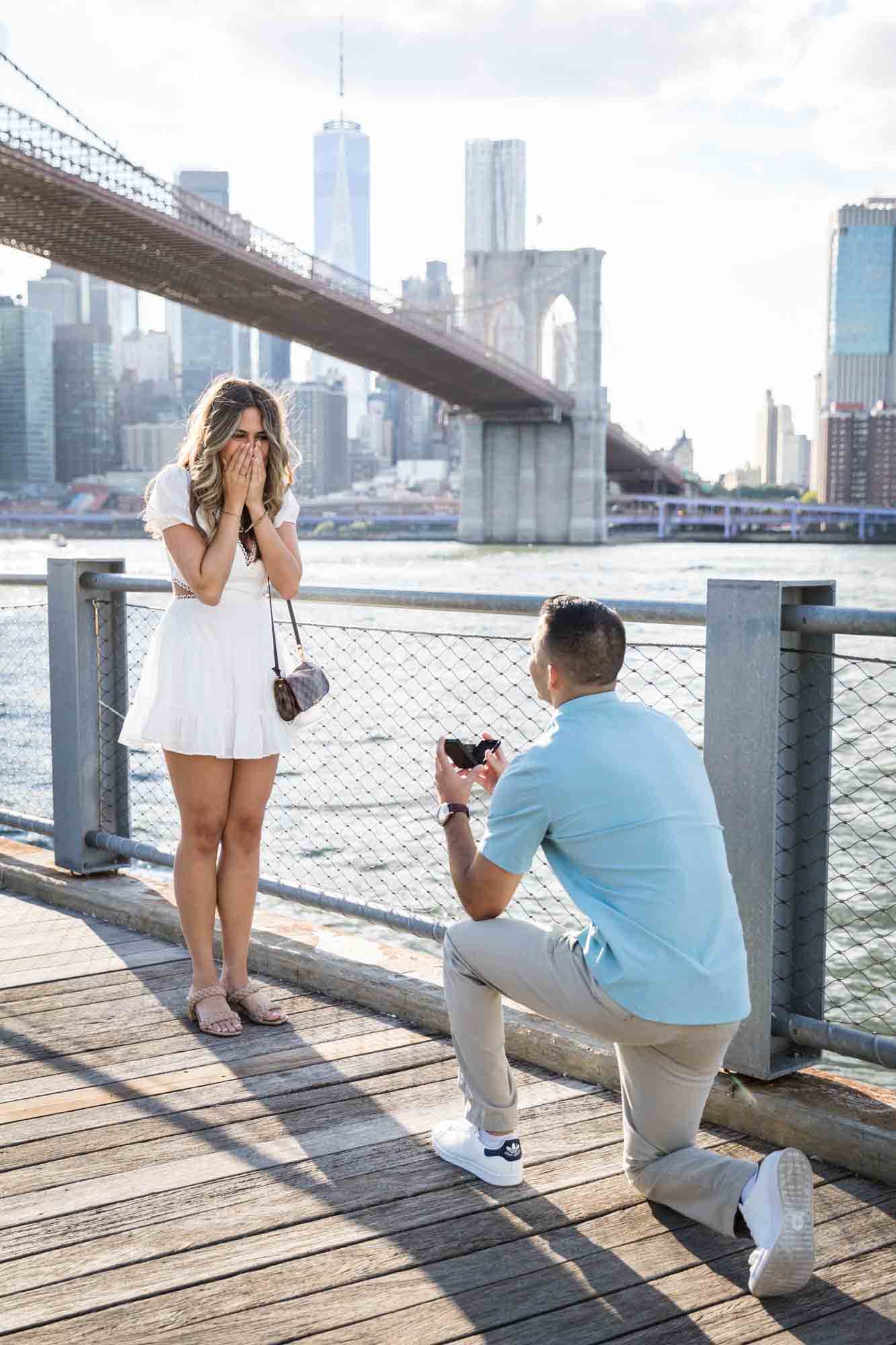 Man on bended knee proposing to woman on boardwalk during a Brooklyn Bridge Park surprise proposal