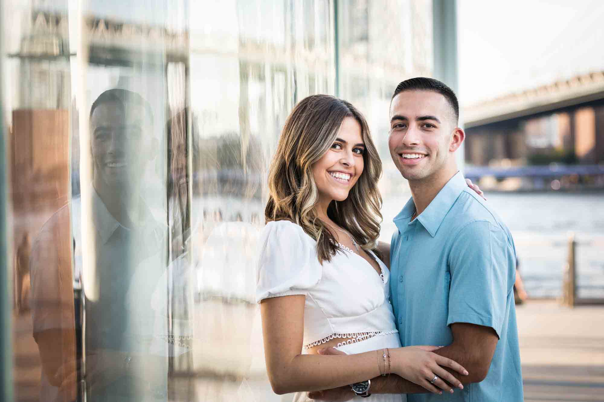 Couple leaning against glass wall of Jane's Carousel during a surprise proposal in Brooklyn Bridge Park