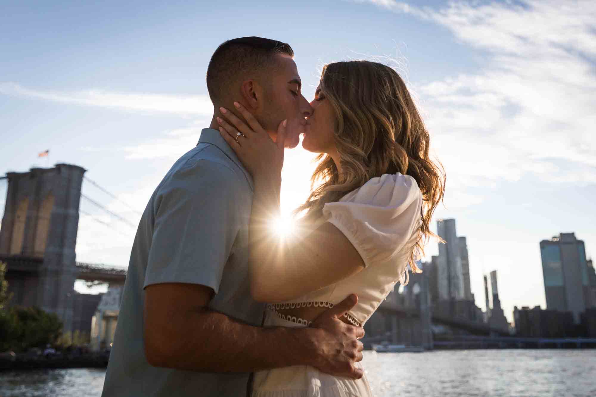 Backlit photo of couple kissing in front of Brooklyn Bridge and waterfront for an article on how to propose in Brooklyn Bridge Park