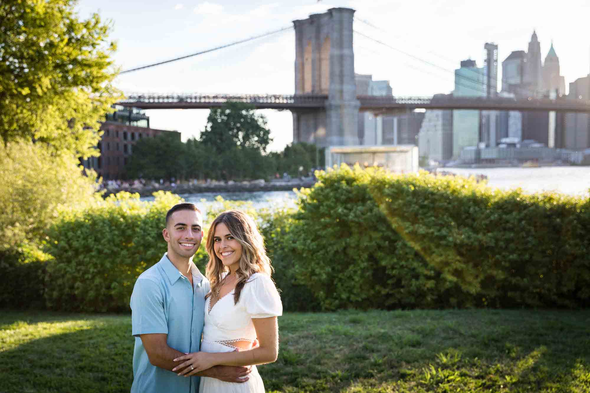 Couple hugging in grass with Brooklyn Bridge and river in background