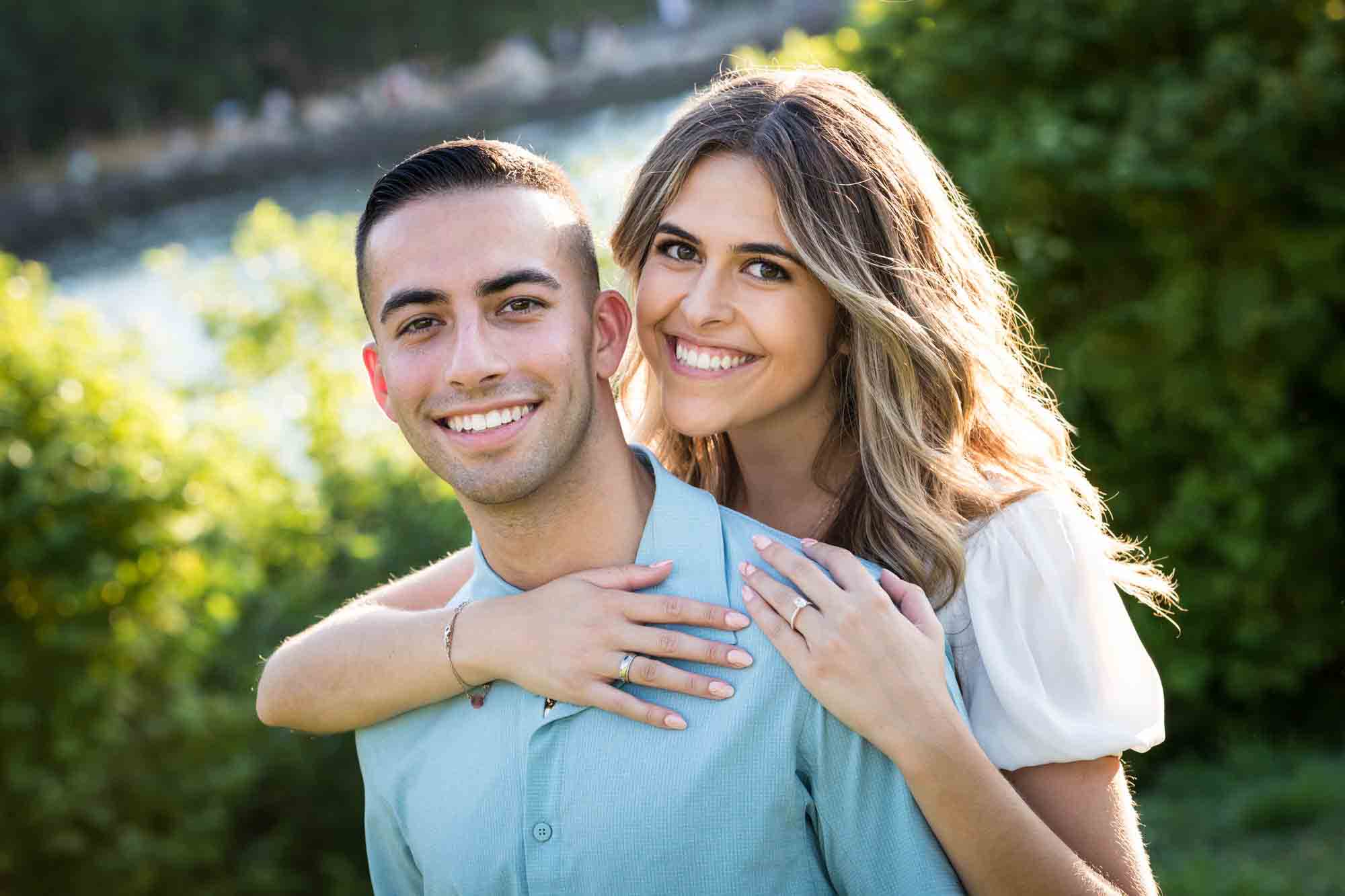 Smiling couple with woman behind man in front of green bushes for an article on how to propose in Brooklyn Bridge Park