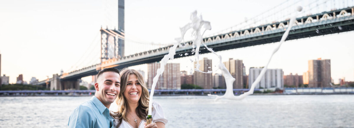 Couple with champagne bottle spewing bubbles in front of Manhattan Bridge for an article on how to propose in Brooklyn Bridge Park