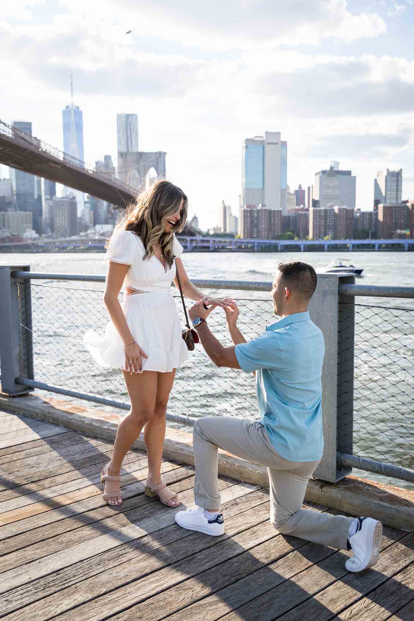 Man on bended knee putting ring on woman's finger during a Brooklyn Bridge Park surprise proposal