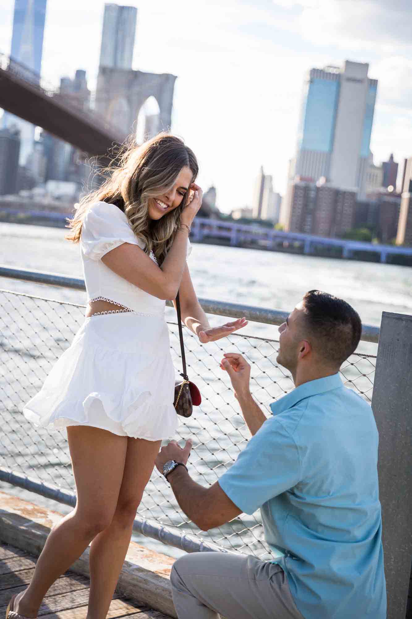 Woman looking down on engagement ring on her hand with man on knee during a Brooklyn Bridge Park surprise proposal