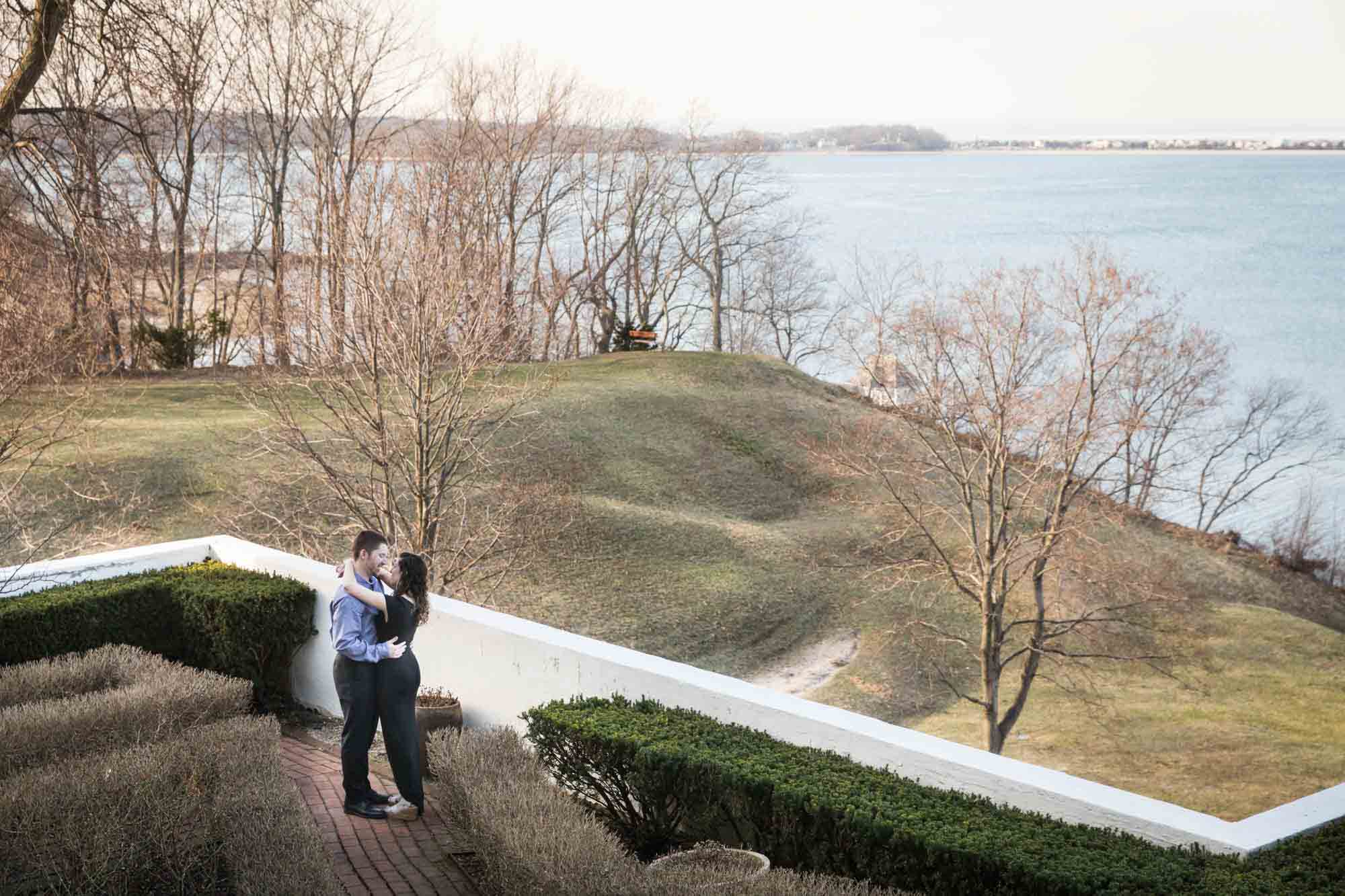 Couple hugging on terrace in front of vista of grass and Northport Bay at a Vanderbilt Museum engagement photo shoot