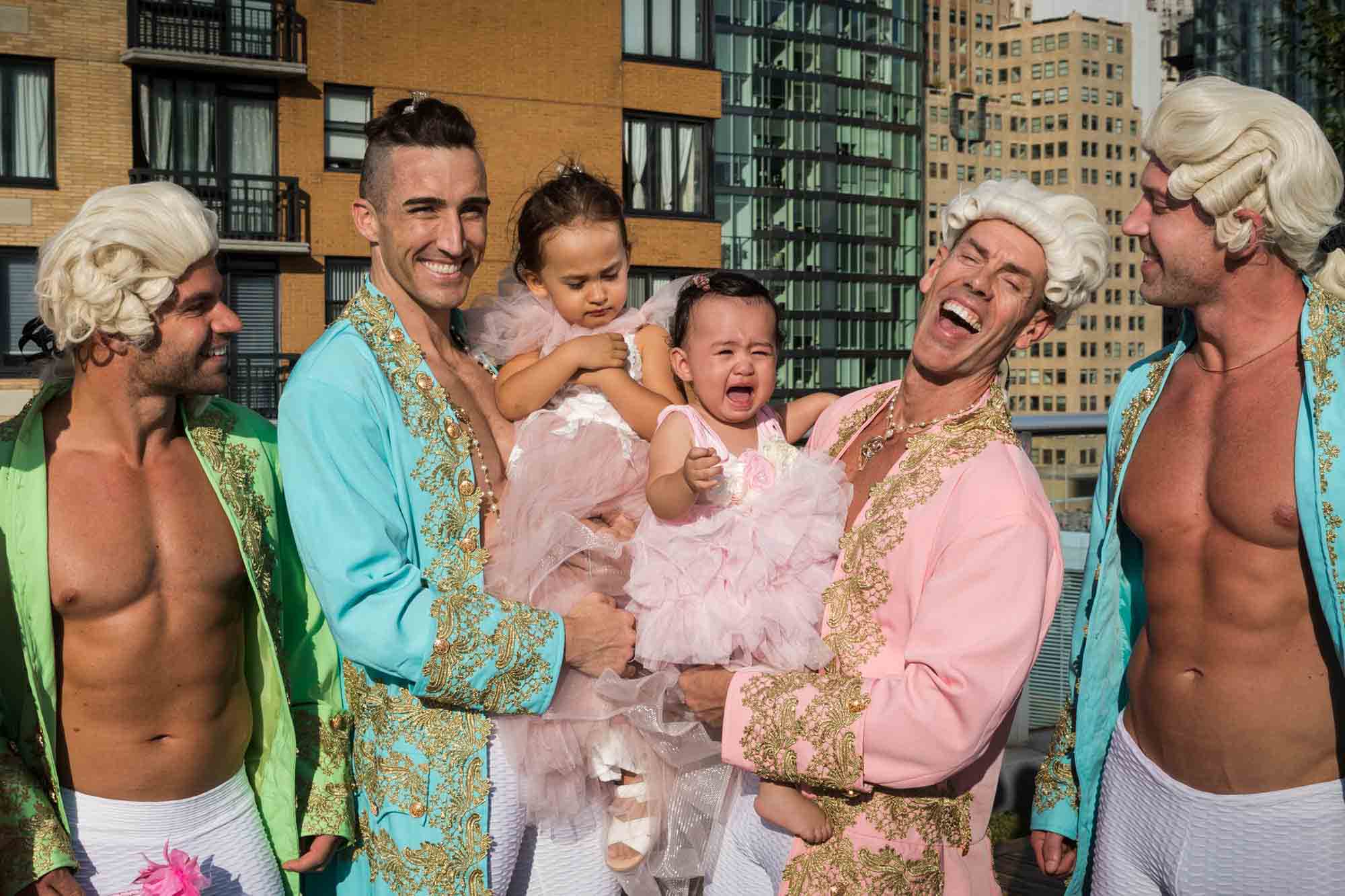 Two men smiling while holding two little girls dressed in pink dresses and crying for an article on event planning photography tips