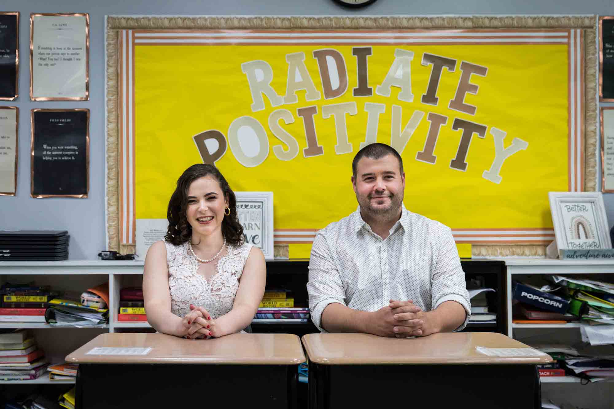Couple sitting at school desks in front of yellow bulletin board for an article on how to recreate your love story in your engagement shoot