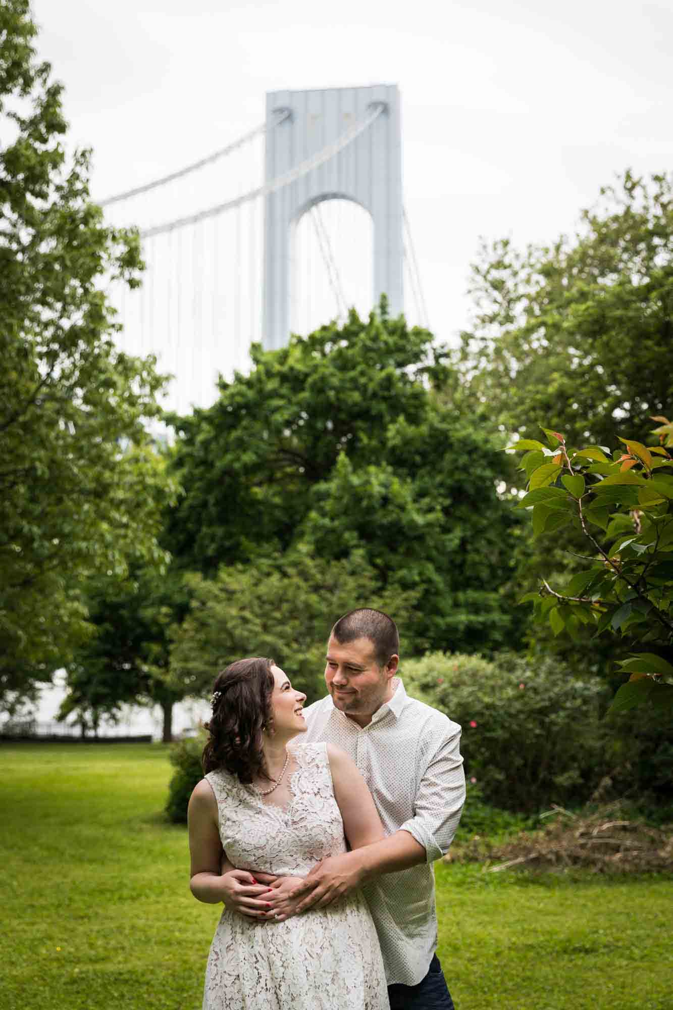 Couple hugging in park with Verrazano Bridge in background during a Bay Ridge engagement shoot